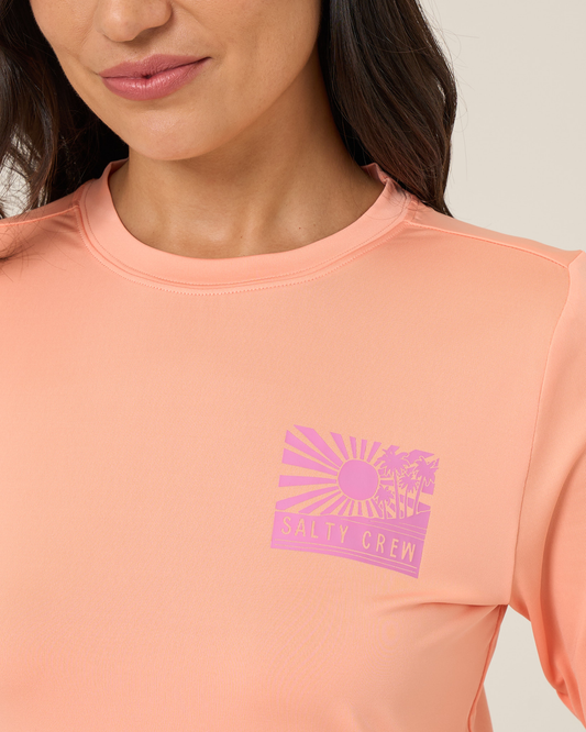 A woman wearing the Salty Crew Rise And Smile Sunshirt in Papaya. This UPF 50+ long-sleeve sunshirt features a pink chest graphic with a sunburst, palm trees, and the text SALTY CREW. Crafted from moisture-wicking fabric, it provides excellent sun protection while keeping you comfortable all day.