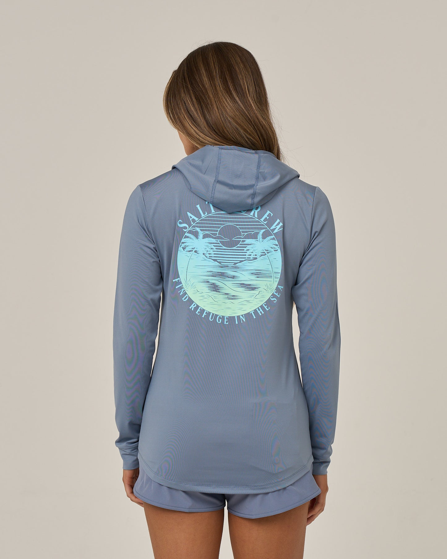 A woman with brown hair, facing away, wears the Salty Crew Tropics Hooded Sunshirt in Flint and shorts. The moisture-wicking shirt features a graphic of a sun over waves along with the words “SALTY CREW FIND REFUGE IN NATURE.”.