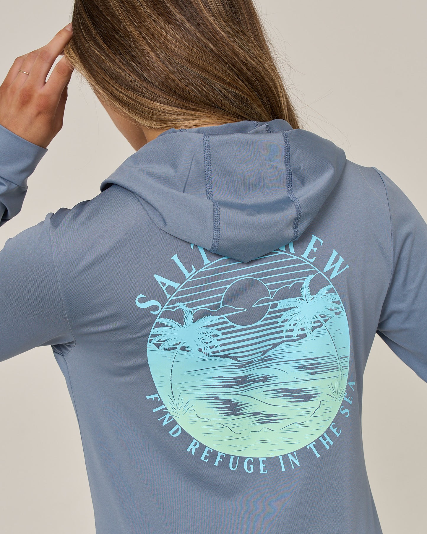 A person wearing the Salty Crew Tropics Hooded Sunshirt in Flint, featuring UPF 50+ protection and a back graphic of a sunset, palm trees, and ocean with the text “Salt New” and “Find Refuge in the Sea,” touches their head with one hand.