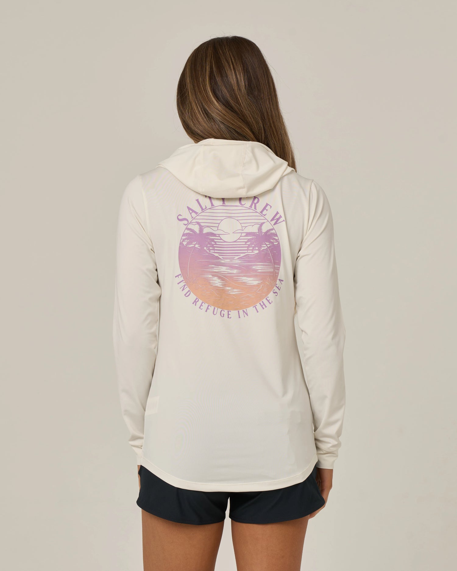 A person with long brown hair wears the Salty Crew Tropics Hooded Sunshirt in Off White, featuring a moisture-wicking design, a pink sunset graphic, and “FIND REFUGE IN THE SEA” text on the back. Paired with dark shorts and set against a plain background, this stylish top functions as a long sleeve sun shirt, providing UPF 50+ protection for comfortable and safe outdoor wear.