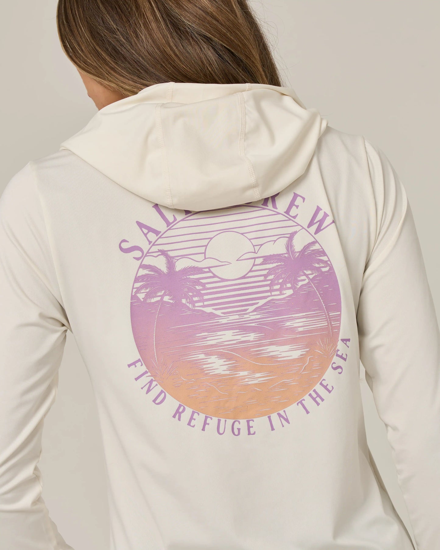 A person wearing the Salty Crew Tropics Hooded Sunshirt in Off White, featuring a pastel sunset graphic of palm trees and ocean. The design includes “Salt Crew” text above and “Find Refuge in the Sea” below the image. This UPF 50+ moisture-wicking shirt delivers both comfort and sun protection for all your seaside adventures.