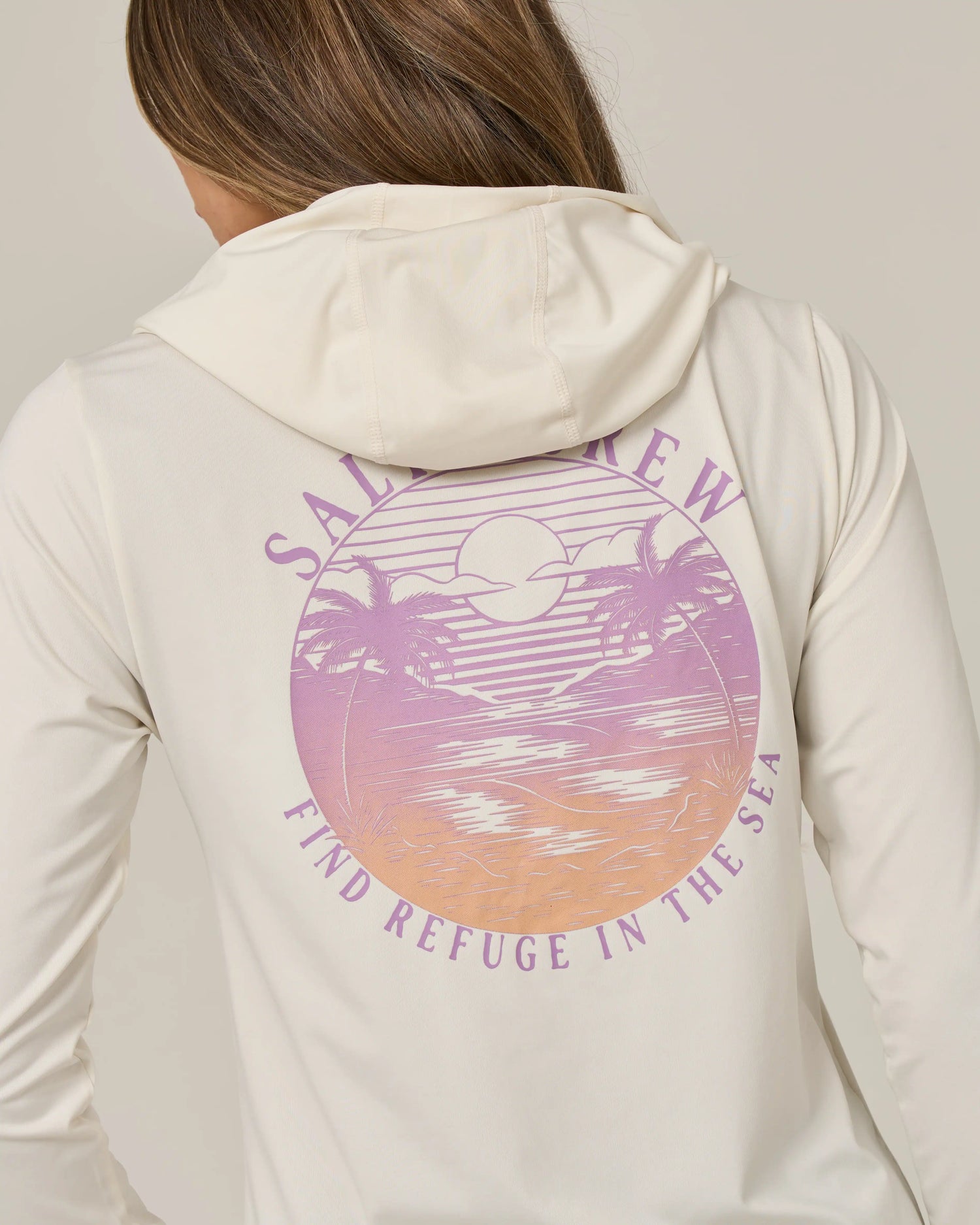 A person wearing the Salty Crew Tropics Hooded Sunshirt in Off White, featuring a pastel sunset graphic of palm trees and ocean. The design includes “Salt Crew” text above and “Find Refuge in the Sea” below the image. This UPF 50+ moisture-wicking shirt delivers both comfort and sun protection for all your seaside adventures.
