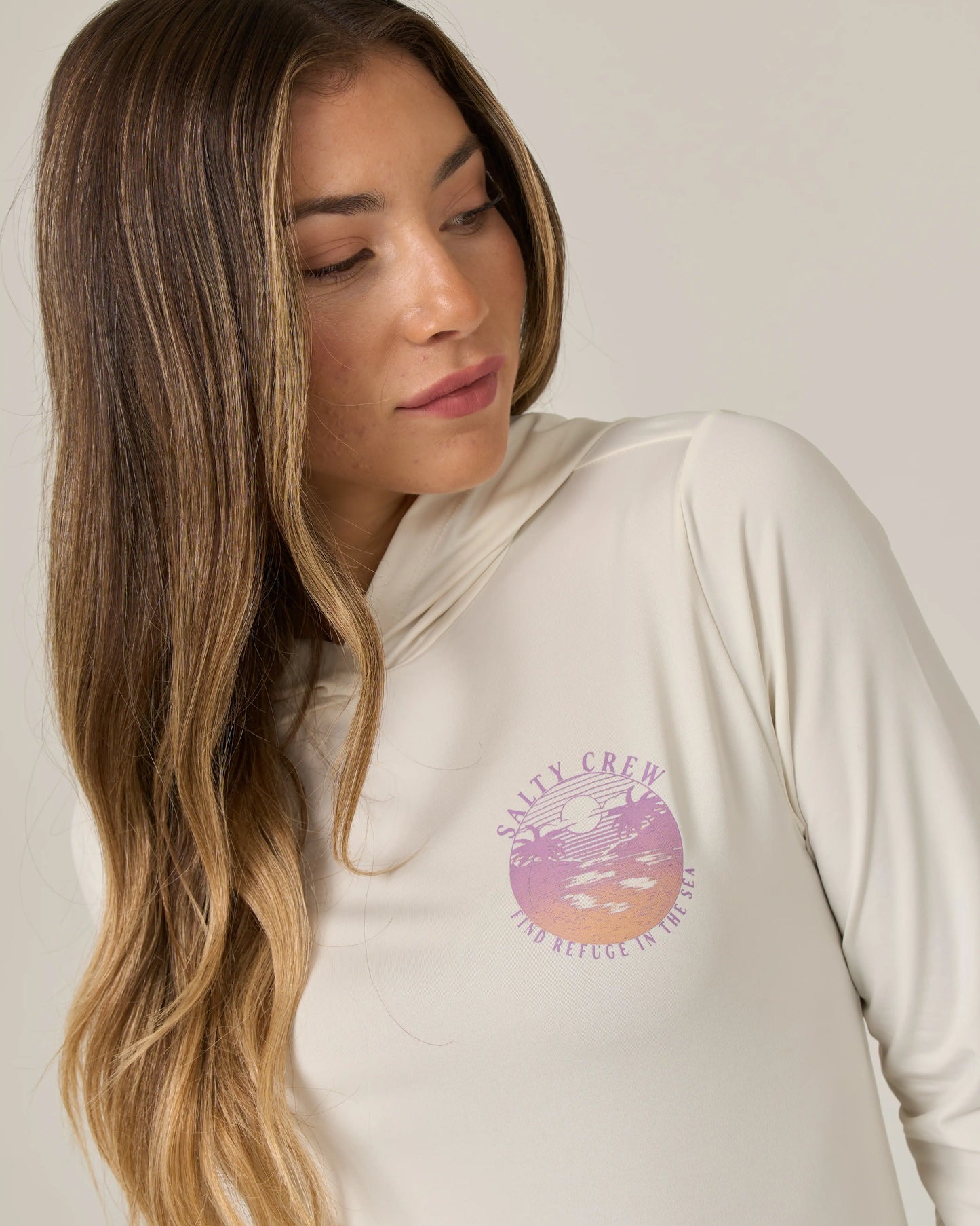An image shows a woman with long brown hair looking down and to the side, wearing the Salty Crew Tropics Hooded Sunshirt in Off White, featuring a round pink graphic and Salty Crew text on the chest.