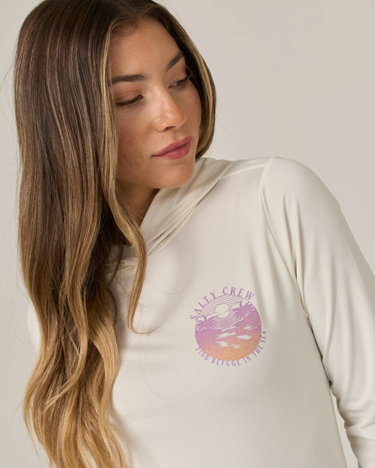 An image shows a woman with long brown hair looking down and to the side, wearing the Salty Crew Tropics Hooded Sunshirt in Off White, featuring a round pink graphic and Salty Crew text on the chest.