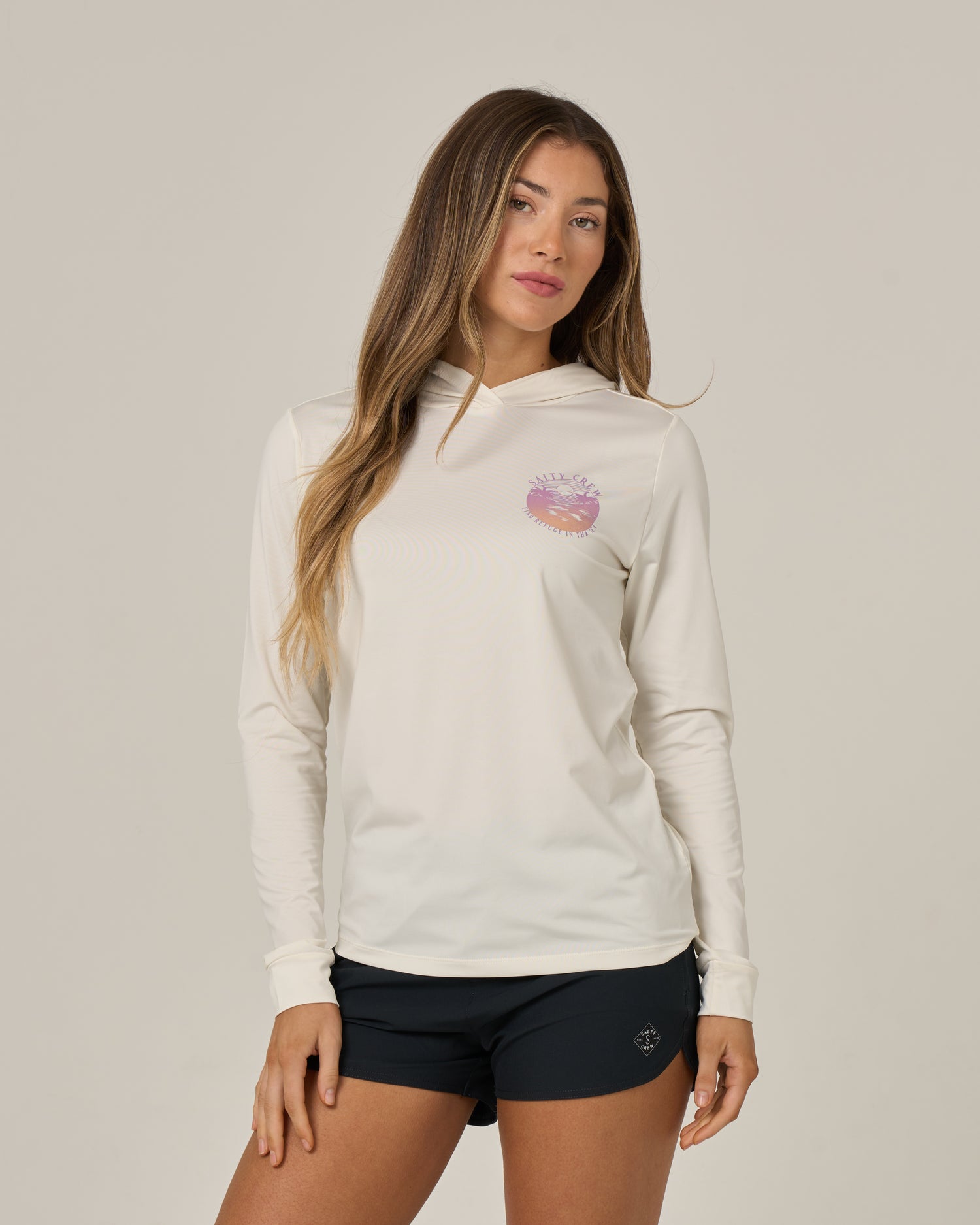 A woman with long hair is wearing the Salty Crew Tropics Hooded Sunshirt in off white, featuring UPF 50+ protection and a pink graphic on the chest, paired with black shorts and standing against a plain light gray background.