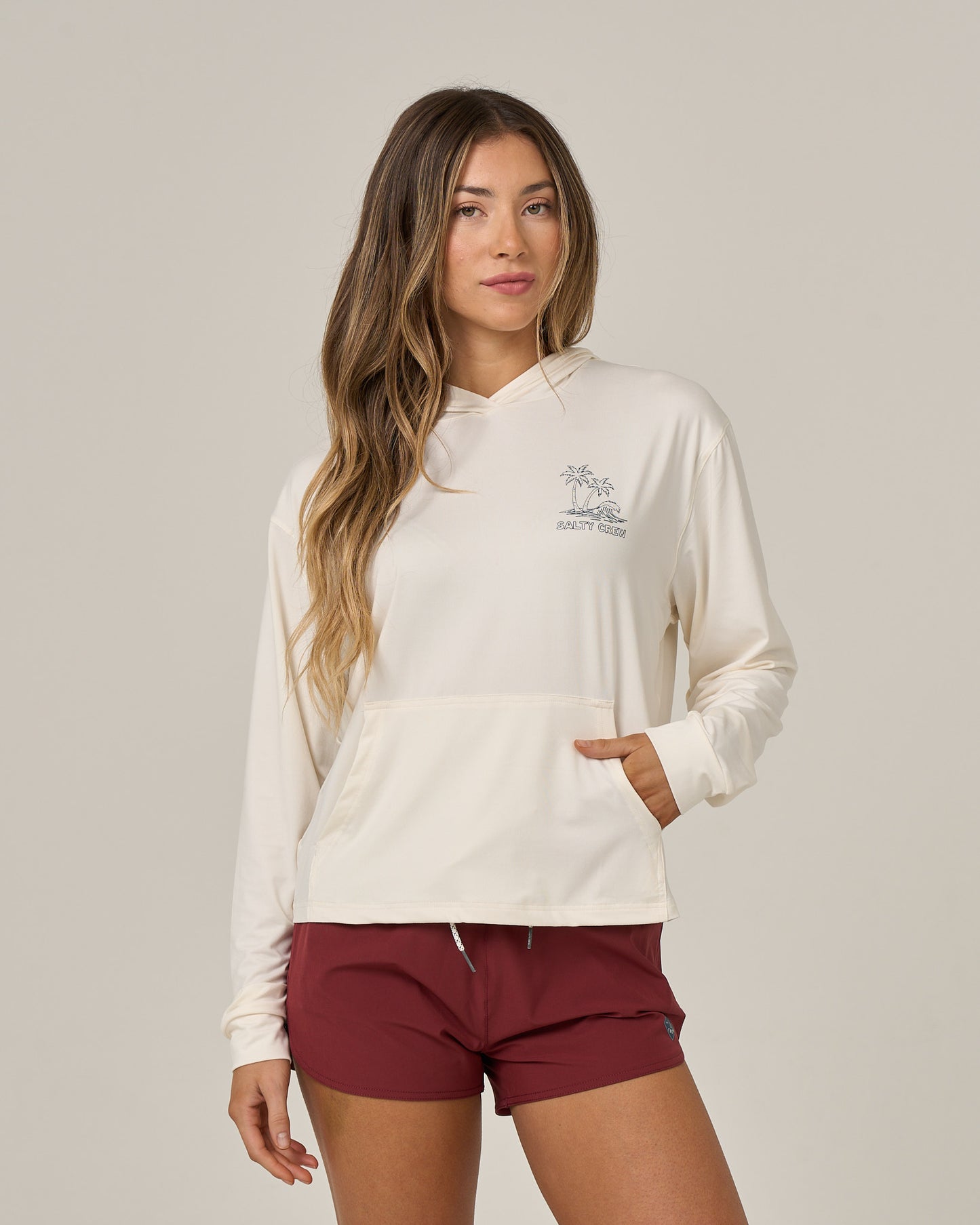 A woman with long brown hair wears the Salty Crew Island Wave Tech Hoodie in Off White, featuring UPF 50+ sun protection and a graphic on the left chest. She pairs it with maroon shorts and stands against a plain light background, one hand in her pocket.