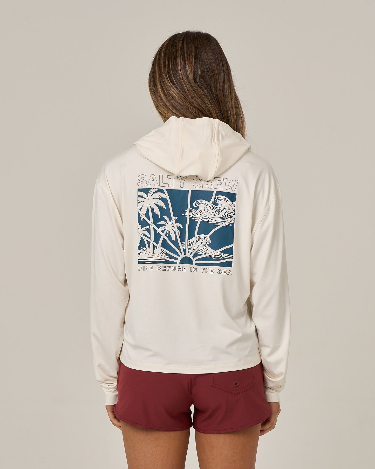 A person is shown from the back against a plain background, wearing the Salty Crew Island Wave Tech Hoodie in Off White—a skimmer length hoody featuring UPF 50+ sun protection and a blue graphic of waves, palm trees, and “SALTY CREW FIND REFUGE IN THE SEA” on the back. The look is paired with maroon shorts.