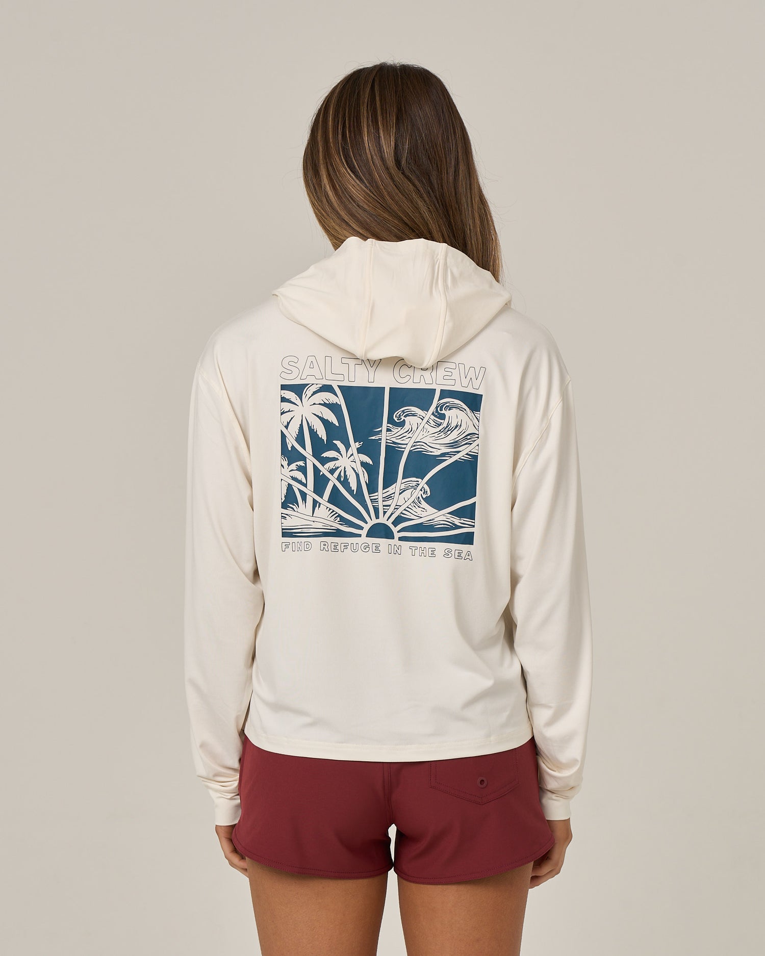 A person is shown from the back against a plain background, wearing the Salty Crew Island Wave Tech Hoodie in Off White—a skimmer length hoody featuring UPF 50+ sun protection and a blue graphic of waves, palm trees, and “SALTY CREW FIND REFUGE IN THE SEA” on the back. The look is paired with maroon shorts.