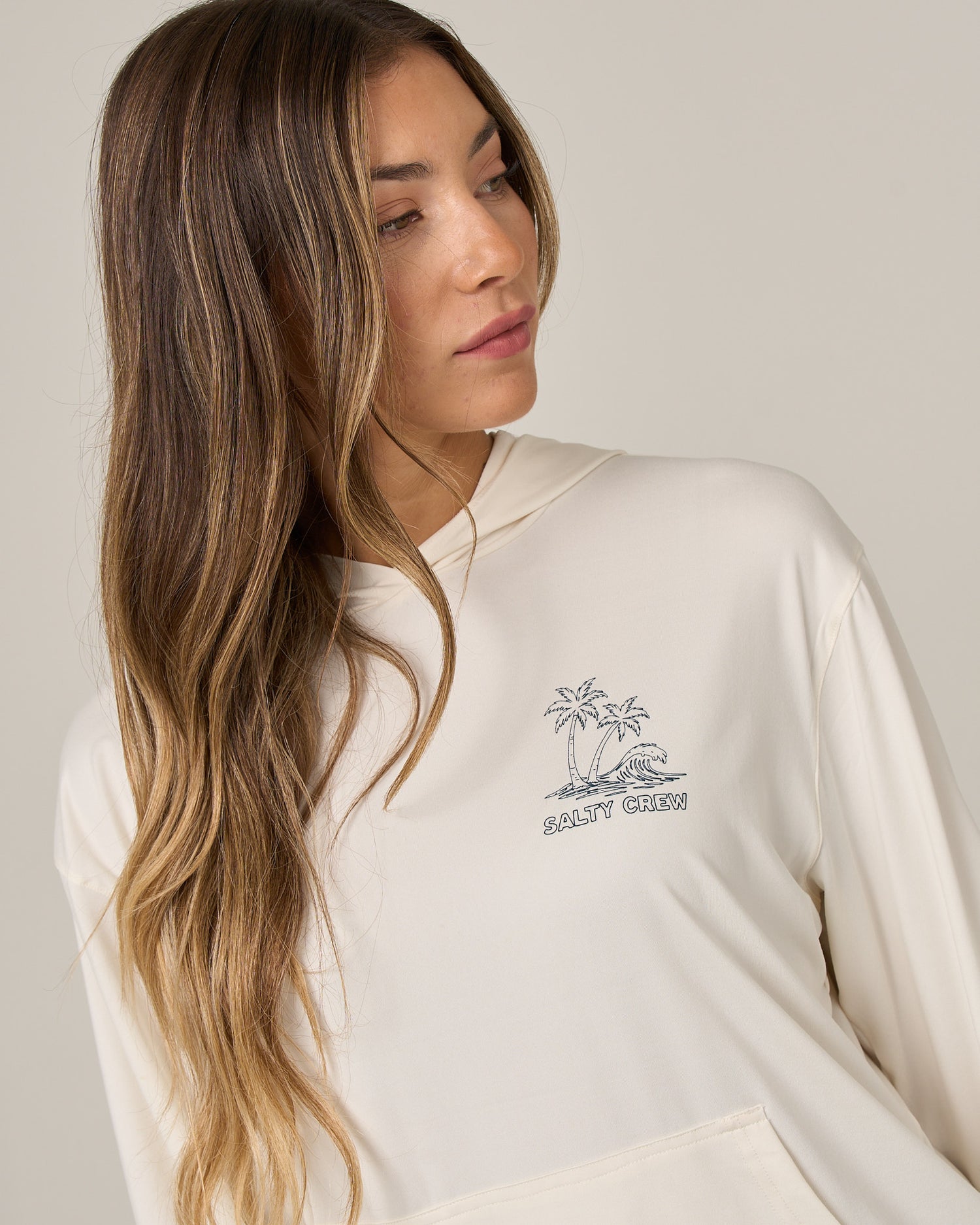 A woman with long, wavy brown hair looks to her left while wearing the Salty Crew Island Wave Tech Hoodie in Off White—a skimmer-length style featuring a small palm tree graphic and “Salty Crew” printed on the front, crafted from lightweight multi-stretch fabric for comfort.