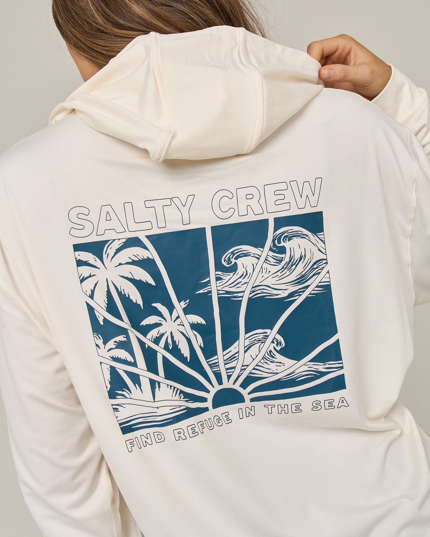 Wearing the Salty Crew Island Wave Tech Hoodie in Off White, which features a skimmer length fit and a blue graphic of palm trees, waves, and sun rays. Designed with lightweight multi-stretch fabric and UPF 50+ sun protection, the hoodie displays “SALTY CREW” at the top and “FIND REFUGE IN THE SEA” at the bottom of the artwork.
