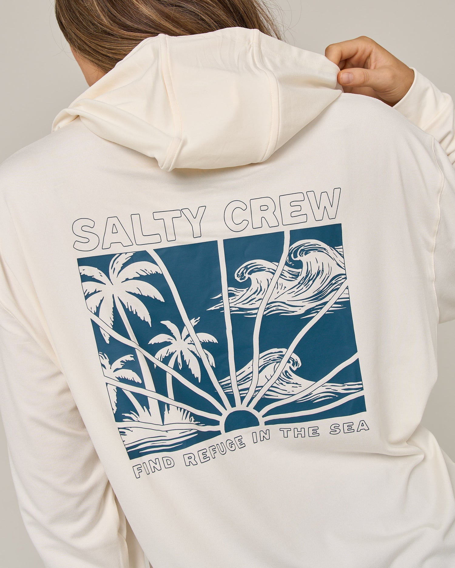 Wearing the Salty Crew Island Wave Tech Hoodie in Off White, which features a skimmer length fit and a blue graphic of palm trees, waves, and sun rays. Designed with lightweight multi-stretch fabric and UPF 50+ sun protection, the hoodie displays “SALTY CREW” at the top and “FIND REFUGE IN THE SEA” at the bottom of the artwork.