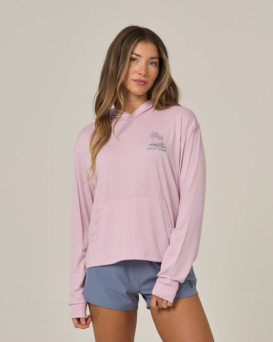 A woman stands against a plain background wearing the Salty Crew Island Wave Tech Hoodie in Pale Orchid, paired with blue athletic shorts, looking at the camera with a relaxed expression.