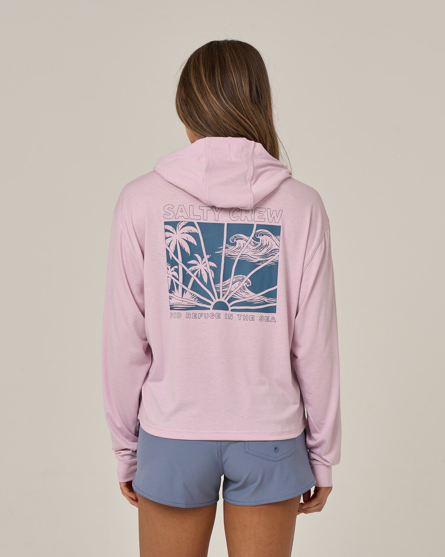 A person stands with their back to the camera against a plain background, wearing the Salty Crew Island Wave Tech Hoodie in Pale Orchid—a lightweight hoody crafted from recycled polyester and featuring a blue graphic of palm trees and birds. Paired with blue shorts, the hoodie provides UPF 50+ sun protection, making it ideal for bright days outdoors.