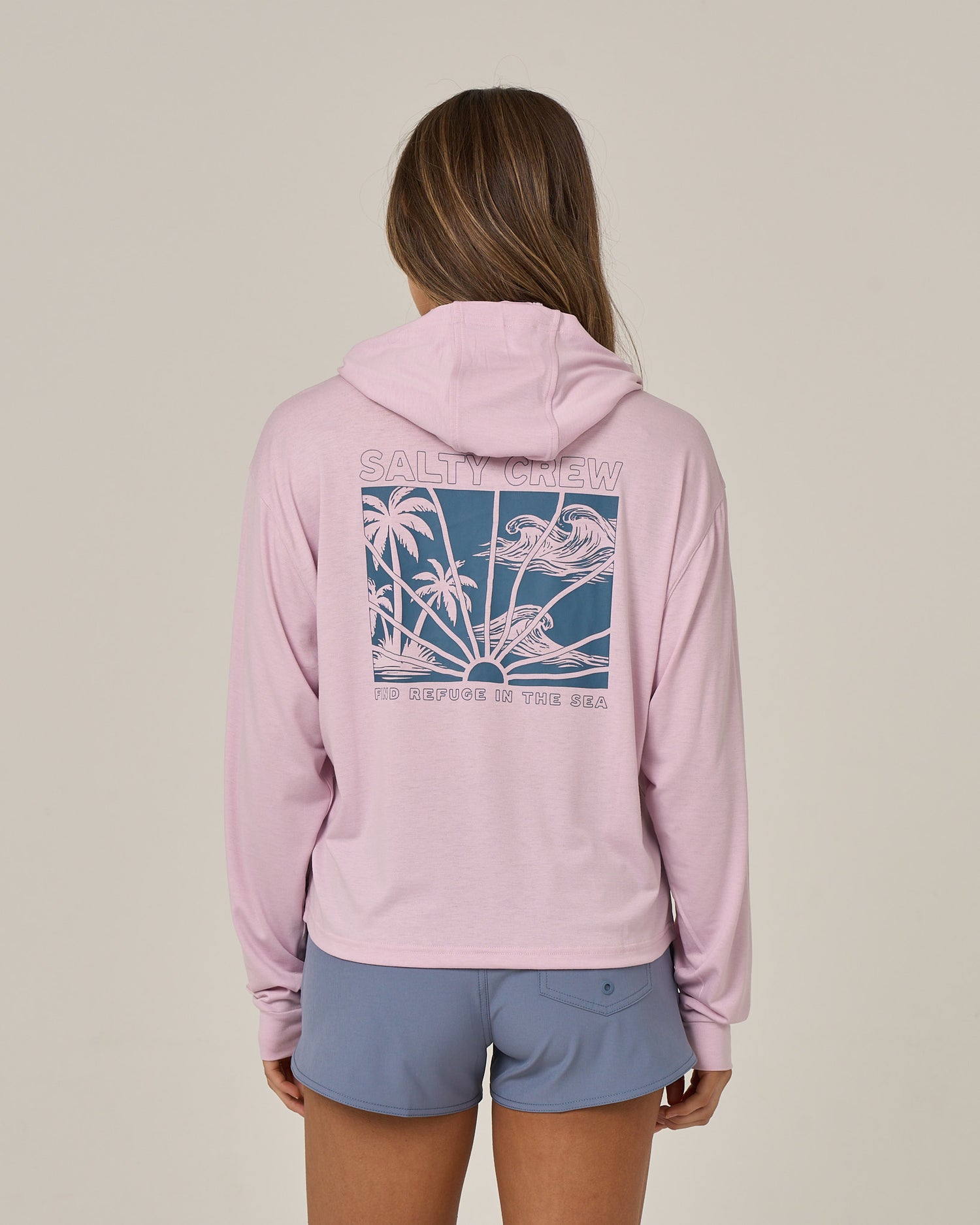 A person stands with their back to the camera against a plain background, wearing the Salty Crew Island Wave Tech Hoodie in Pale Orchid—a lightweight hoody crafted from recycled polyester and featuring a blue graphic of palm trees and birds. Paired with blue shorts, the hoodie provides UPF 50+ sun protection, making it ideal for bright days outdoors.