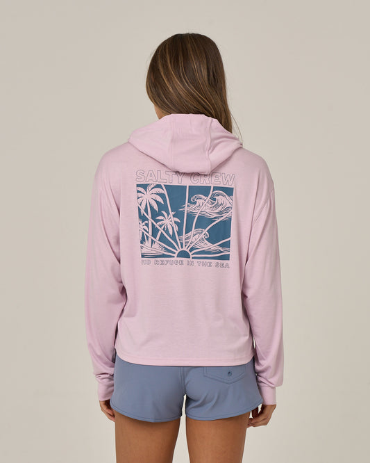 A person stands with their back to the camera against a plain background, wearing the Salty Crew Island Wave Tech Hoodie in Pale Orchid—a lightweight hoody crafted from recycled polyester and featuring a blue graphic of palm trees and birds. Paired with blue shorts, the hoodie provides UPF 50+ sun protection, making it ideal for bright days outdoors.