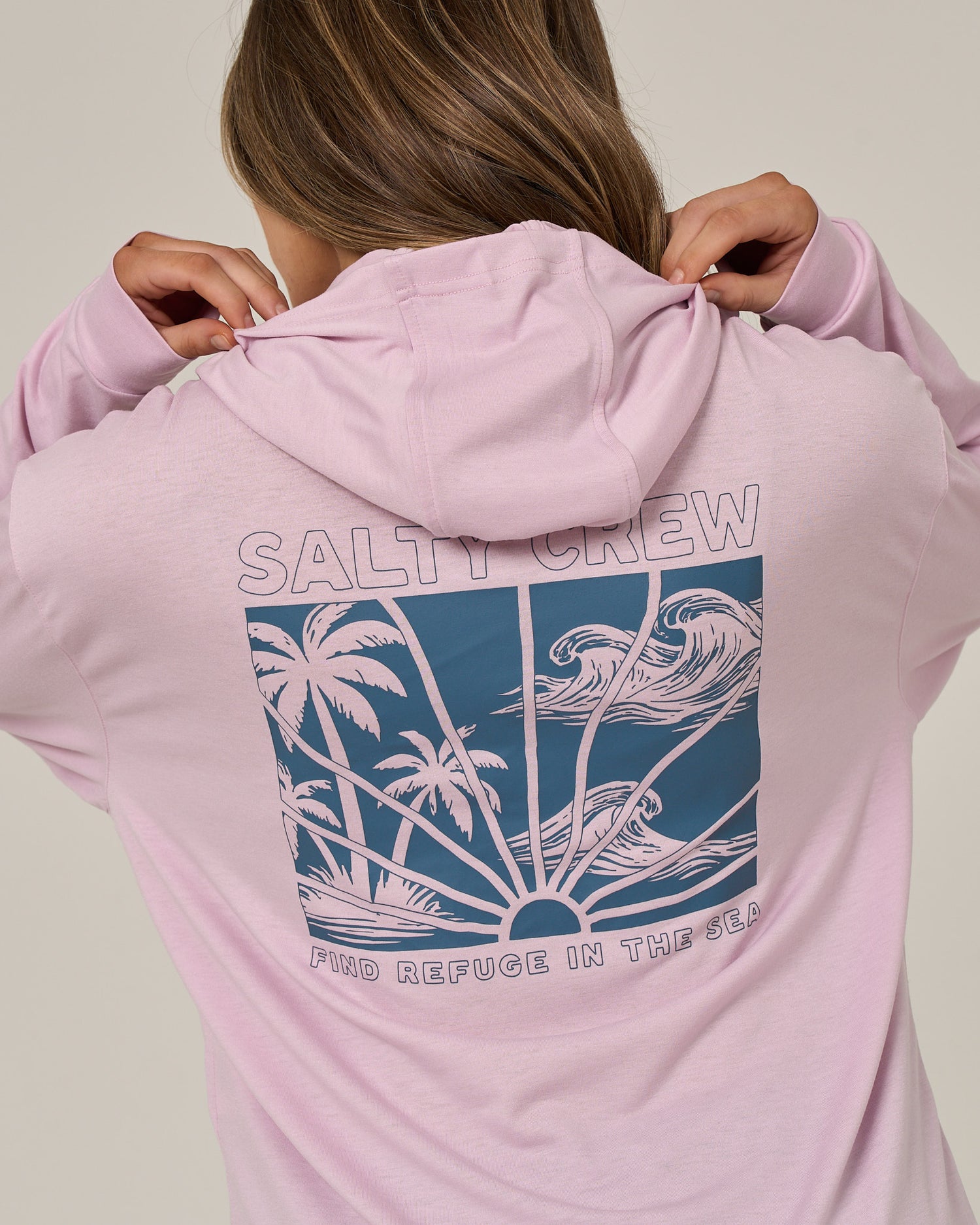A person wears the Salty Crew Island Wave Tech Hoodie in Pale Orchid, a lightweight hoody crafted from recycled polyester and offering UPF 50+ sun protection. The back features a graphic of palm trees, waves, and the sun, with “SALTY CREW” and “FIND REFUGE IN THE SEA” printed on it. The person is pulling up the hood.