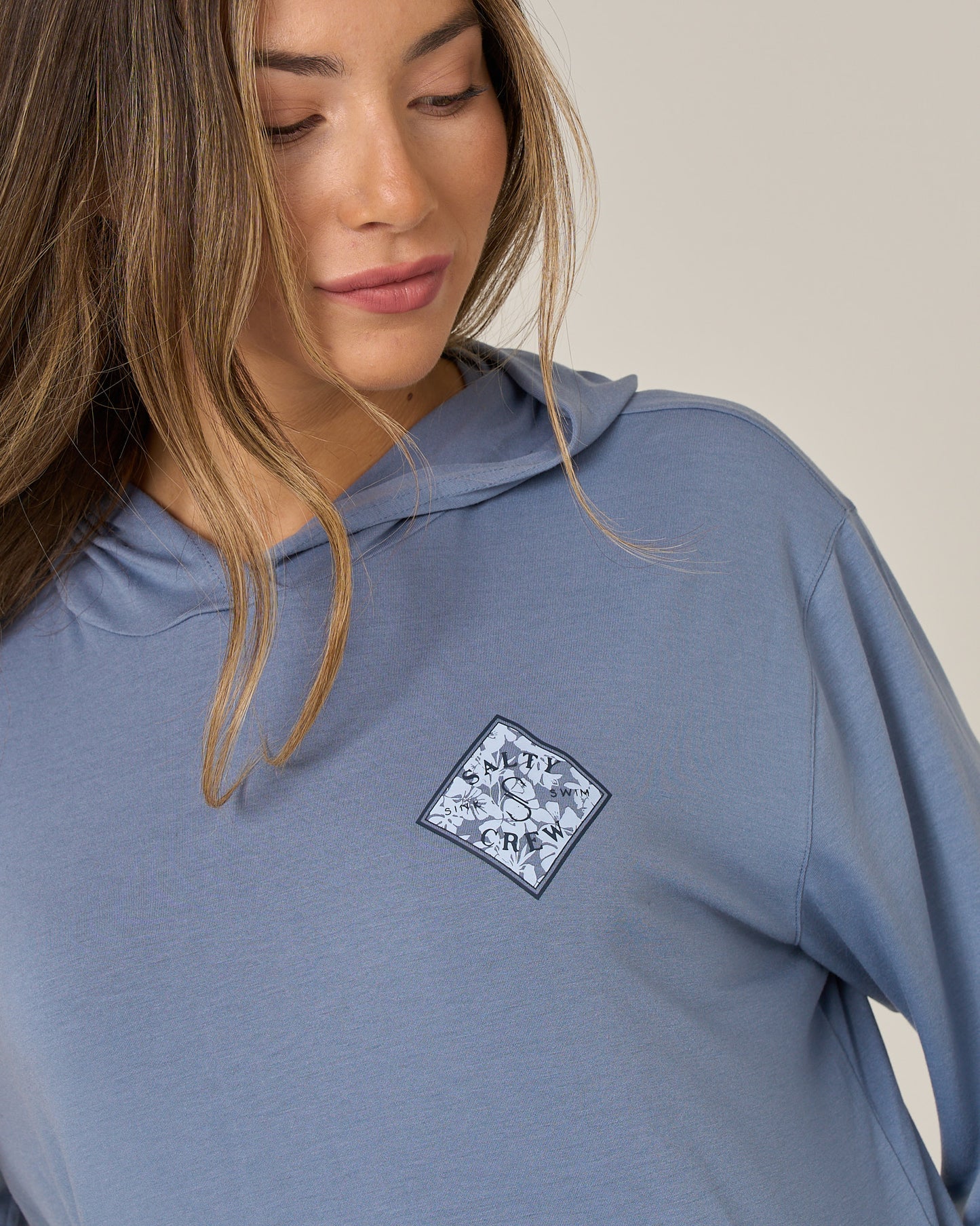 A woman with long brown hair wears the Salty Crew Tippet Fill Tech Hoodie in Flint, a lightweight, light blue hoody featuring a diamond-shaped patch with a wave design and the letters S, W, T, Y on the chest. Made from recycled polyester with UPF 50+ sun protection, she looks down and smiles softly.