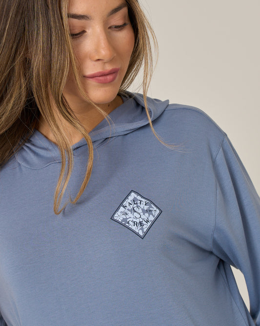 A woman with long brown hair wears the Salty Crew Tippet Fill Tech Hoodie in Flint, a lightweight, light blue hoody featuring a diamond-shaped patch with a wave design and the letters S, W, T, Y on the chest. Made from recycled polyester with UPF 50+ sun protection, she looks down and smiles softly.