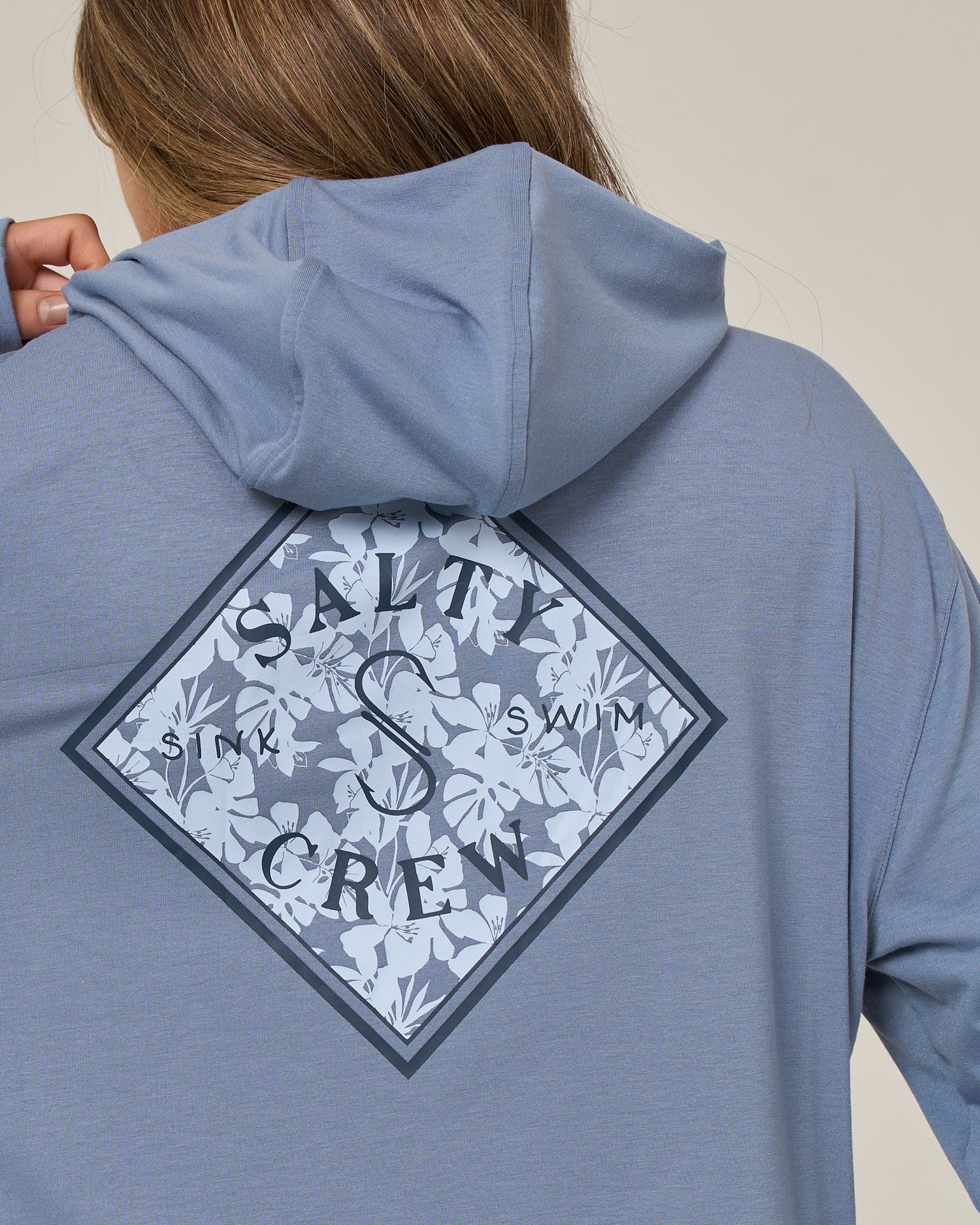 A person is seen from behind wearing the Salty Crew Tippet Fill Tech Hoodie in Flint, a lightweight hoodie made from recycled polyester. The hoodie features a blue color and displays a diamond-shaped floral graphic with the words “SALTY CREW” and “SINK SWIM” printed on the back.