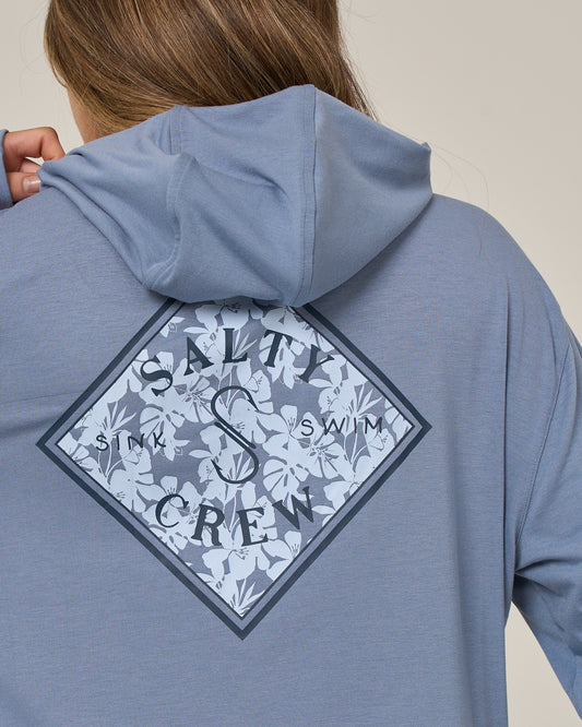 A person is seen from behind wearing the Salty Crew Tippet Fill Tech Hoodie in Flint, a lightweight hoodie made from recycled polyester. The hoodie features a blue color and displays a diamond-shaped floral graphic with the words “SALTY CREW” and “SINK SWIM” printed on the back.