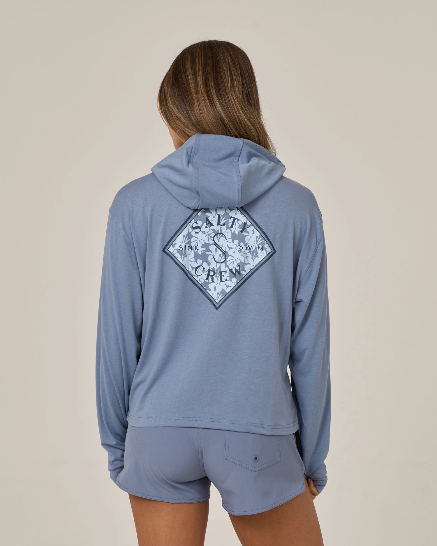 A person with long hair, seen from the back, is wearing the Salty Crew Tippet Fill Tech Hoodie in Flint—a light blue, lightweight hoody crafted from recycled polyester for sustainability. The hoodie showcases a diamond-shaped Salty Crew logo with a floral design on the back and is paired with shorts.
