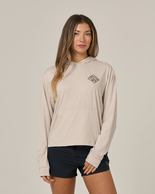 A woman wearing the Salty Crew Tippet Fill Tech Hoodie in Oatmeal, a lightweight hoody made from recycled polyester and featuring a small chest graphic, stands in front of a plain light background, looking at the camera with a neutral expression. The hoodie offers UPF 50+ sun protection for added comfort and safety and is paired with black shorts.
