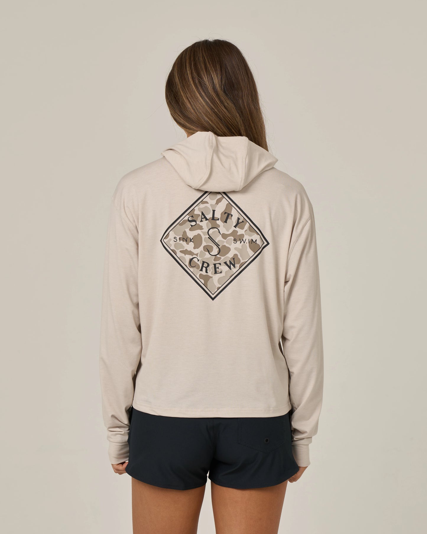 Wearing black shorts and standing with their back to the camera against a plain background, a person with long hair sports the Salty Crew Tippet Fill Tech Hoodie in Oatmeal—a lightweight hoody made from recycled polyester, featuring a Salty Crew logo on the back and offering UPF 50+ sun protection.