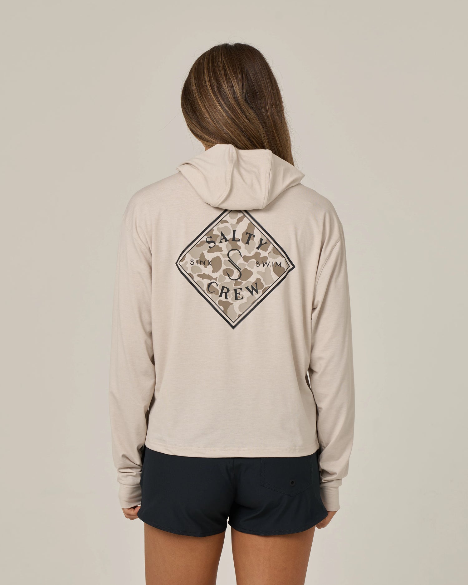 Wearing black shorts and standing with their back to the camera against a plain background, a person with long hair sports the Salty Crew Tippet Fill Tech Hoodie in Oatmeal—a lightweight hoody made from recycled polyester, featuring a Salty Crew logo on the back and offering UPF 50+ sun protection.