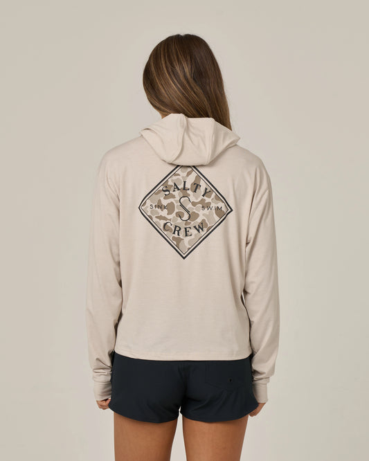 Wearing black shorts and standing with their back to the camera against a plain background, a person with long hair sports the Salty Crew Tippet Fill Tech Hoodie in Oatmeal—a lightweight hoody made from recycled polyester, featuring a Salty Crew logo on the back and offering UPF 50+ sun protection.