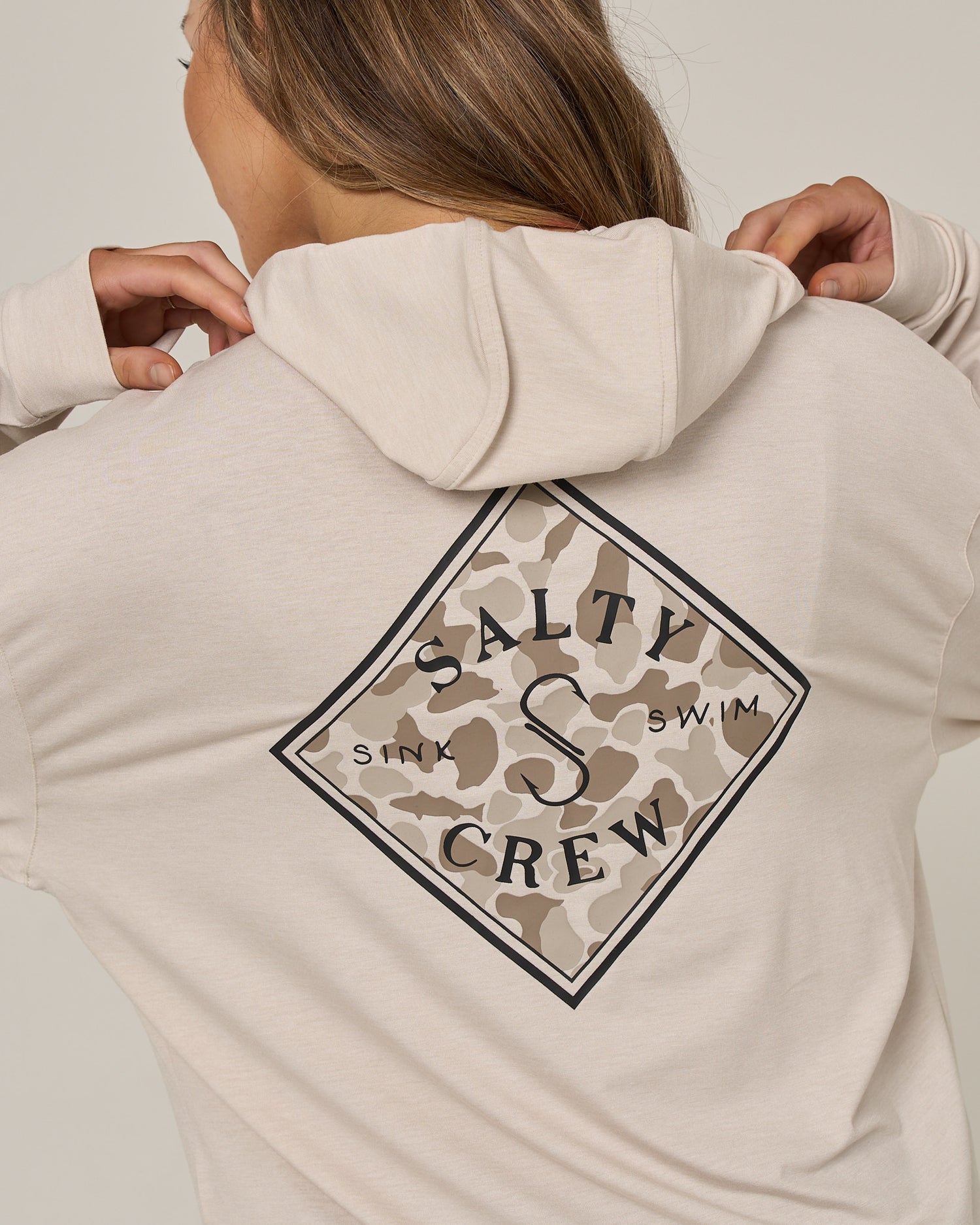 A person wearing the Salty Crew Tippet Fill Tech Hoodie in oatmeal—a light beige, lightweight hoody made from recycled polyester, featuring a camouflage diamond logo on the back that reads “Salty Crew Sink Swim” and offering UPF 50+ sun protection.