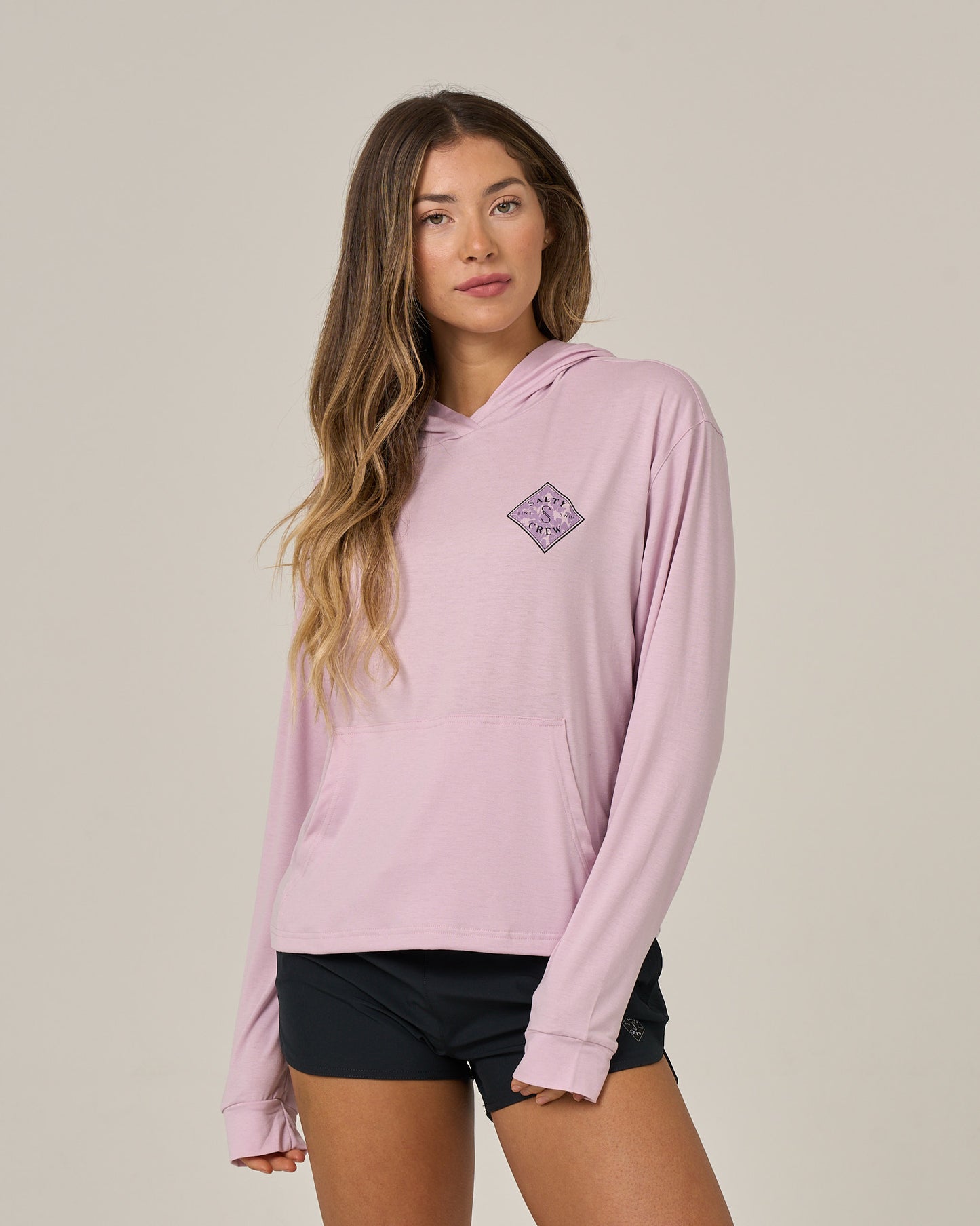 A woman with long, wavy hair wears the Salty Crew Tippet Fill Tech Hoodie in Pale Orchid—a skimmer length hoody featuring a small purple graphic on the chest—paired with black shorts, standing against a plain beige background. Made from lightweight multi-stretch fabric, this hoodie offers comfort and easy movement for everyday wear.