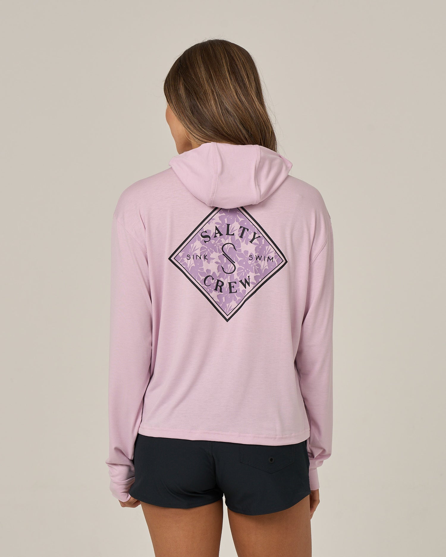 A person with long hair, facing away from the camera against a plain background, is wearing the Salty Crew Tippet Fill Tech Hoodie in Pale Orchid—a skimmer-length hoody showcasing a decorative back design. Paired with black shorts, this lightweight multi-stretch hoodie offers UPF 50+ sun protection.