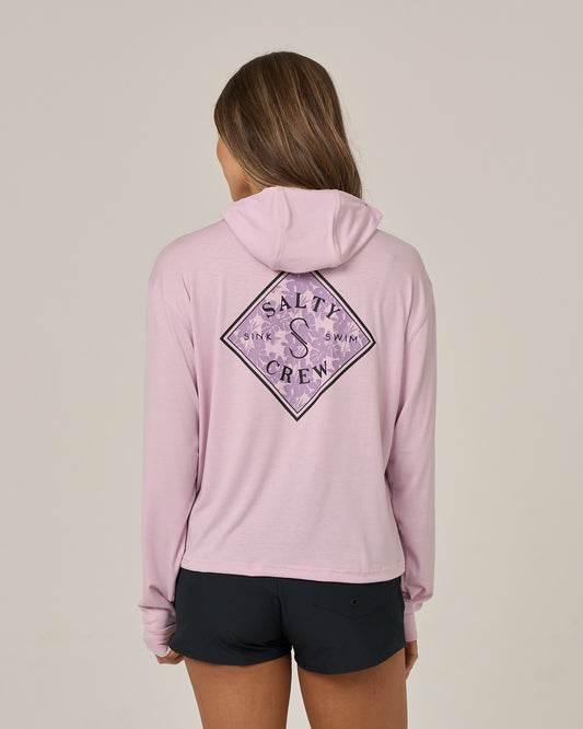 A person with long hair, facing away from the camera against a plain background, is wearing the Salty Crew Tippet Fill Tech Hoodie in Pale Orchid—a skimmer-length hoody showcasing a decorative back design. Paired with black shorts, this lightweight multi-stretch hoodie offers UPF 50+ sun protection.