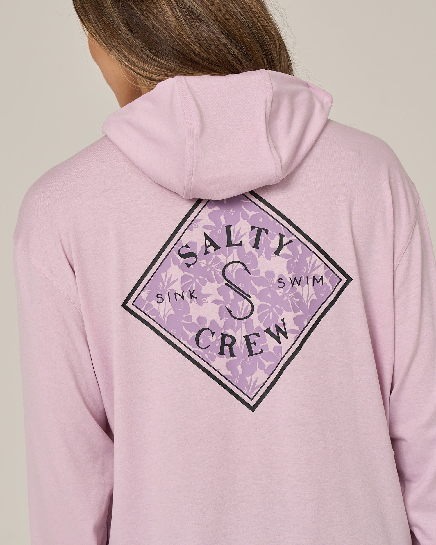 A person is seen from behind wearing the Salty Crew Tippet Fill Tech Hoodie in Pale Orchid. This skimmer-length hoodie features a purple diamond Salty Crew Sink Swim logo on the back, set over a subtle floral pattern. Made from lightweight multi-stretch fabric for comfort and offering UPF 50+ sun protection, it’s perfect for sunny days.