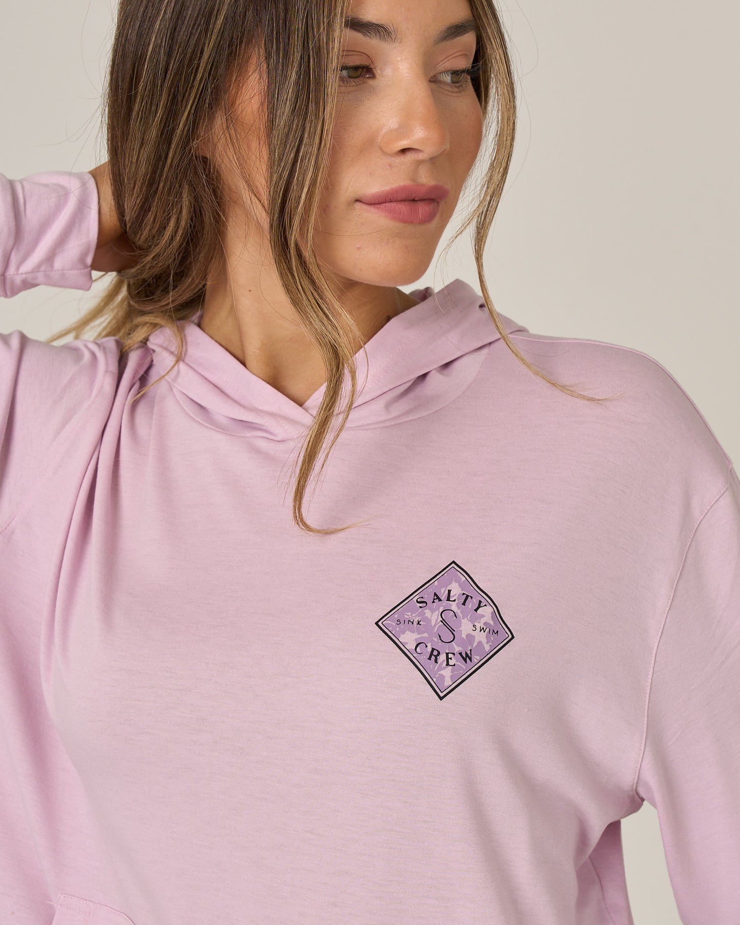 A woman wears the Salty Crew Tippet Fill Tech Hoodie in Pale Orchid, a skimmer-length hoodie featuring the Salty Crew logo on the chest, UPF 50+ sun protection, and lightweight multi-stretch fabric; she looks to her left with one hand behind her head.
