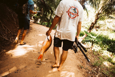 Man wearing Salty Crew T-Shirt walks in jungle holding fish and fishing rods. 