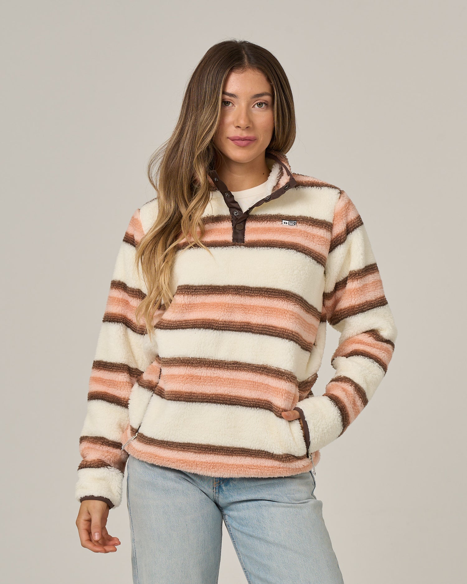 A woman wearing the Salty Crew Calm Seas Pullover in Dusty Coral—a plush, mock neck pullover with brown and orange horizontal stripes—paired with light blue jeans, stands against a plain gray background.