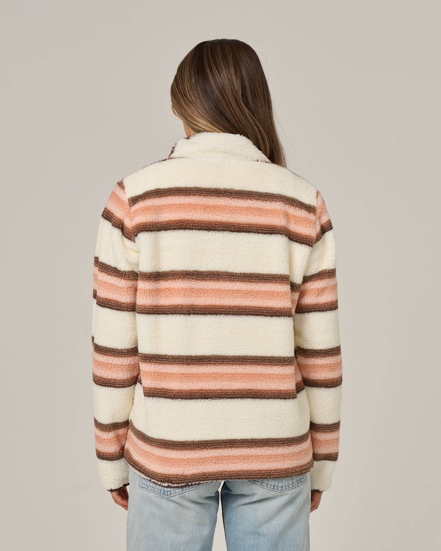 A person with long brown hair, seen from the back, wears the Salty Crew Calm Seas Pullover in Dusty Coral—a cream-colored sherpa jacket featuring horizontal brown and pink stripes and a mock neck collar—paired with light blue jeans against a plain background.