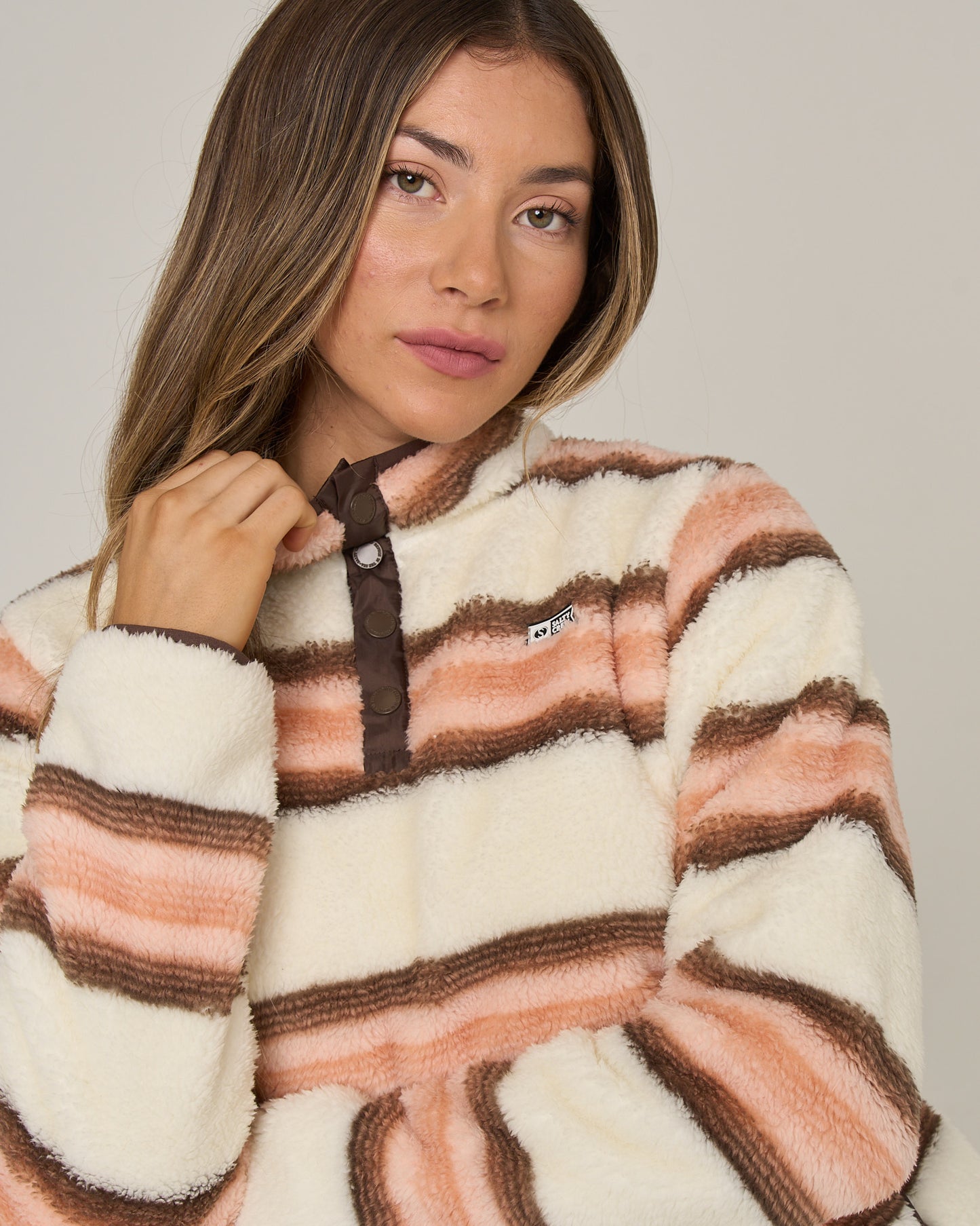 A woman with long brown hair wears the Salty Crew Calm Seas Pullover – Dusty Coral, a plush top with peach, cream, and brown stripes and a cozy mock neck collar. She stands against a plain background and looks at the camera.
