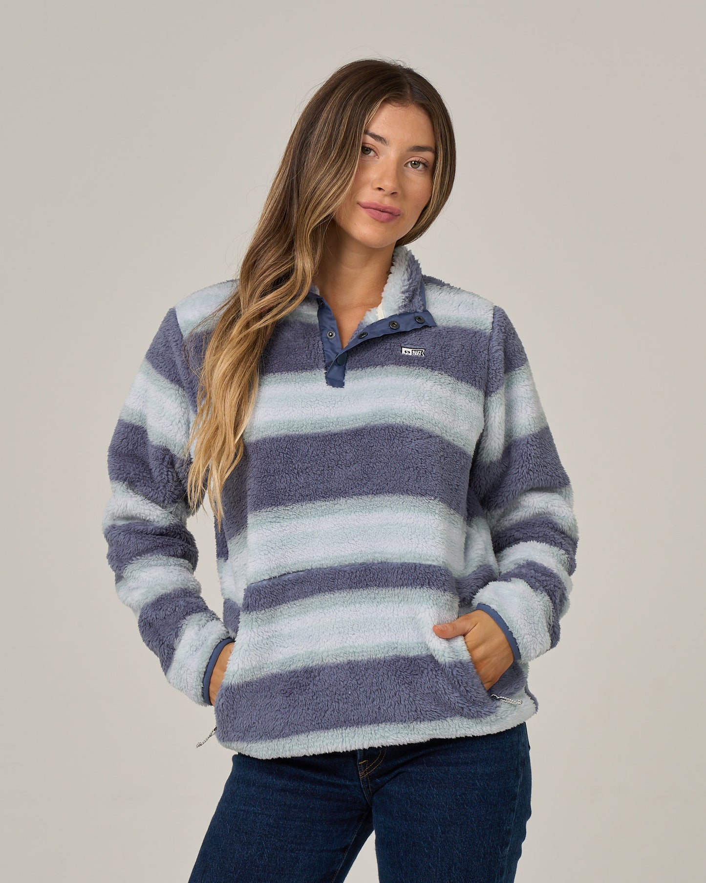 A woman with long, wavy hair stands against a plain background, hands in her pockets, wearing the Salty Crew Calm Seas Pullover in Flint—a plush blue and white striped sweater with a mock neck collar—paired with blue jeans.