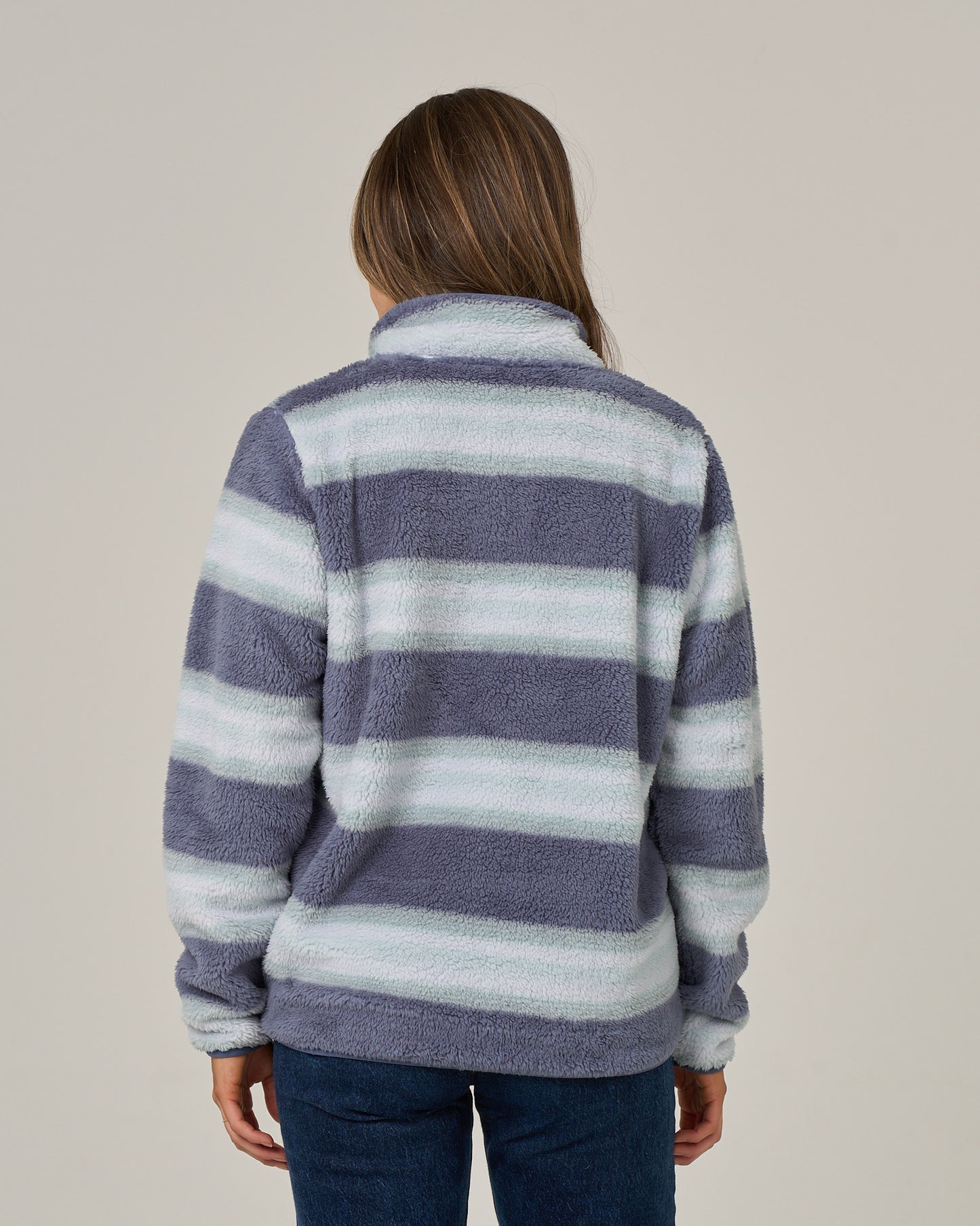 A person with long brown hair is wearing the Salty Crew Calm Seas Pullover in Flint, a sherpa-style jacket in gray and white with fuzzy striped textures and a mock neck collar, paired with blue jeans. The individual is facing away from the camera against a plain background.