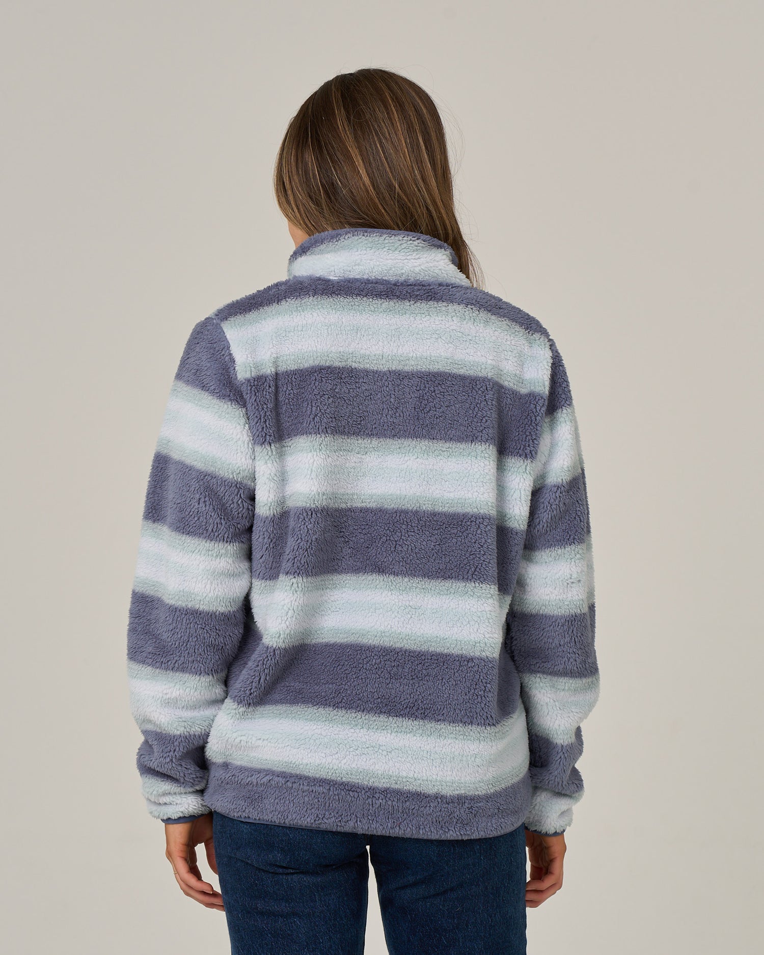 A person with long brown hair is wearing the Salty Crew Calm Seas Pullover in Flint, a sherpa-style jacket in gray and white with fuzzy striped textures and a mock neck collar, paired with blue jeans. The individual is facing away from the camera against a plain background.
