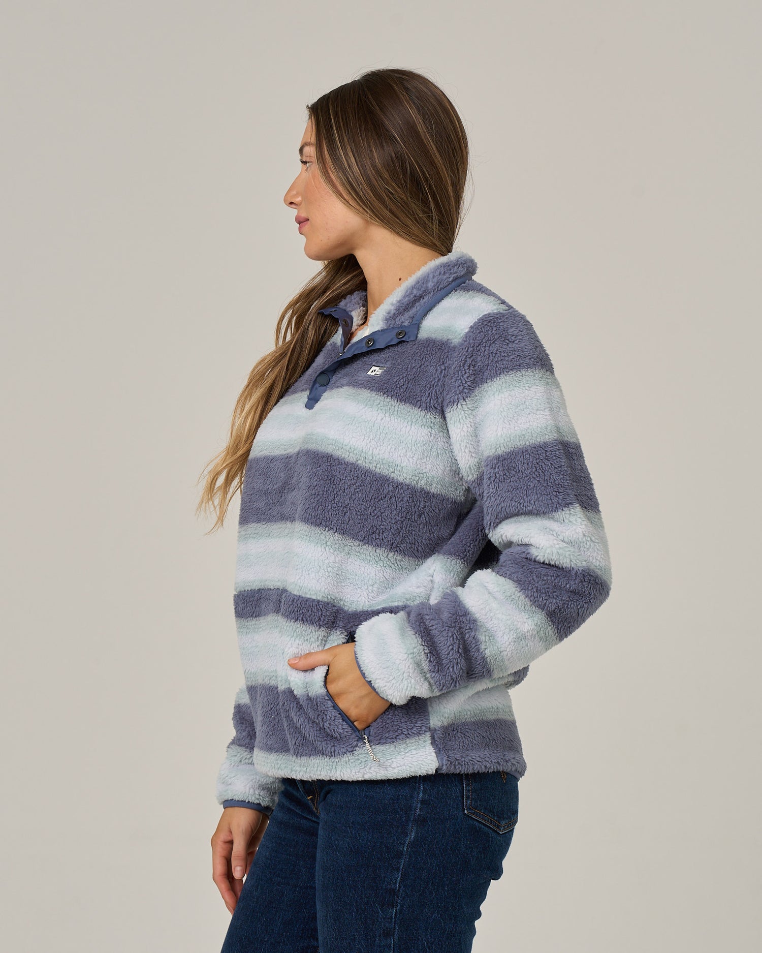 A woman with long brown hair wears the Salty Crew Calm Seas Pullover in Flint—a plush, striped blue and white top featuring a mock neck collar—paired with blue jeans. She stands sideways with one hand in her pocket, looking to her left against a plain light background.