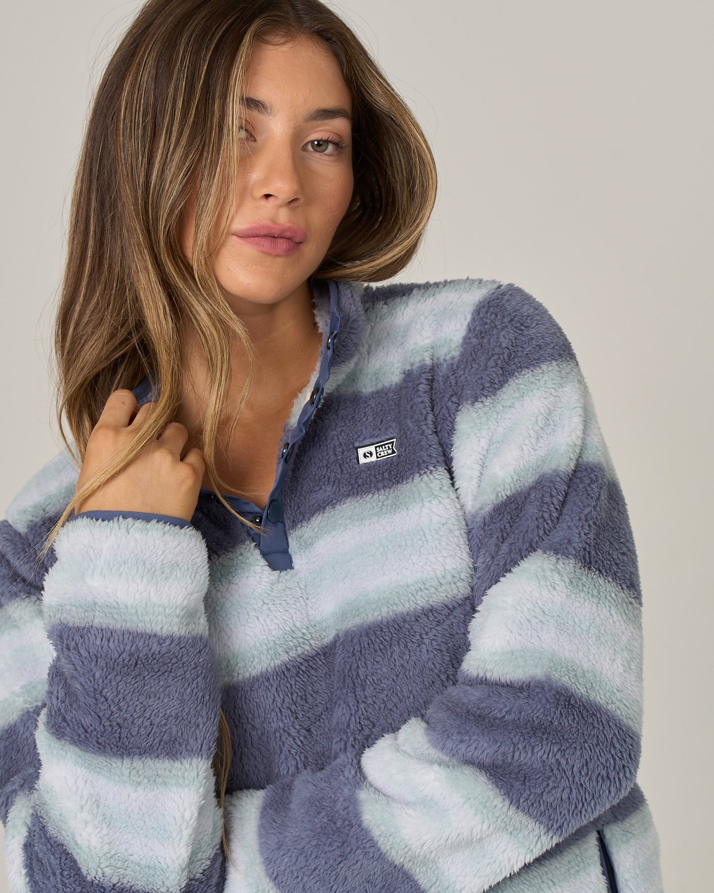 A woman with long hair wears the Salty Crew Calm Seas Pullover in Flint, a soft and plush blue-and-white striped top. Featuring a cozy mock neck collar, she has a relaxed expression as she lightly holds the collar with one hand. The background is plain and neutral.