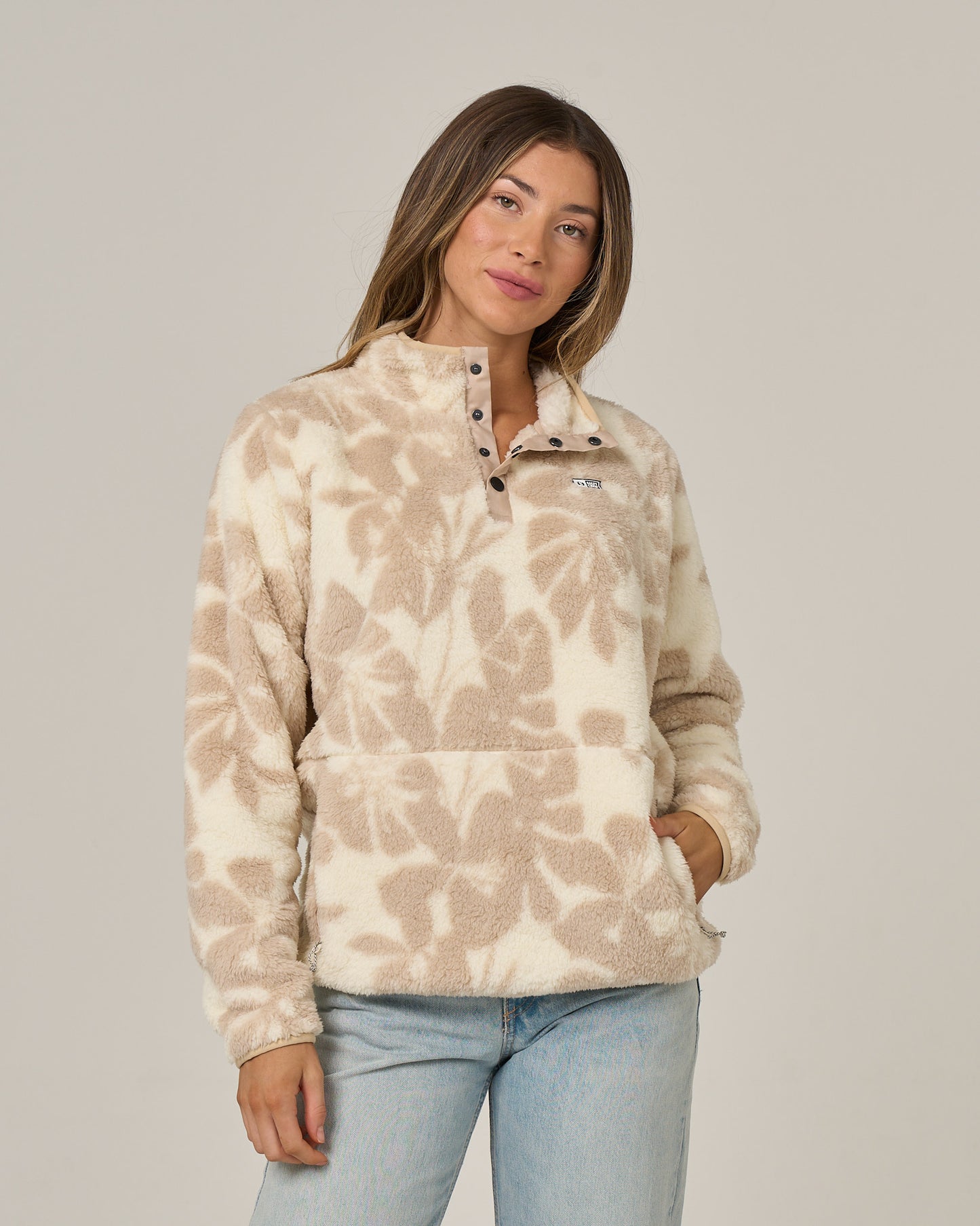 A woman wears the Salty Crew Calm Seas Pullover in Natural—a beige and white patterned sherpa featuring buttons at the mock neck—paired with light blue jeans, standing against a plain background with one hand in her pocket.