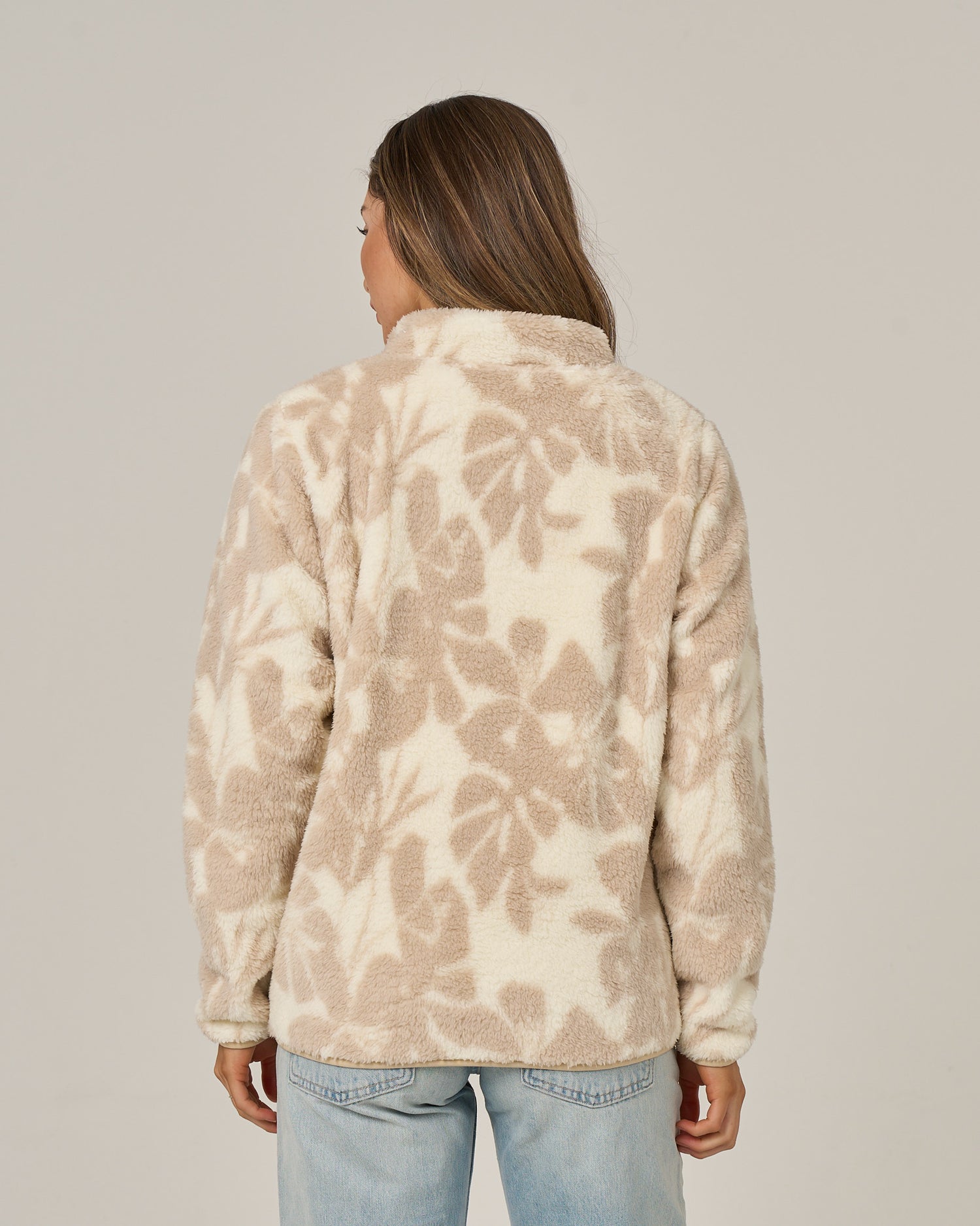 A person with long brown hair, seen from behind, wears the Salty Crew Calm Seas Pullover in Natural—a beige and cream floral sherpa jacket with a mock neck—paired with light blue jeans against a plain light background.