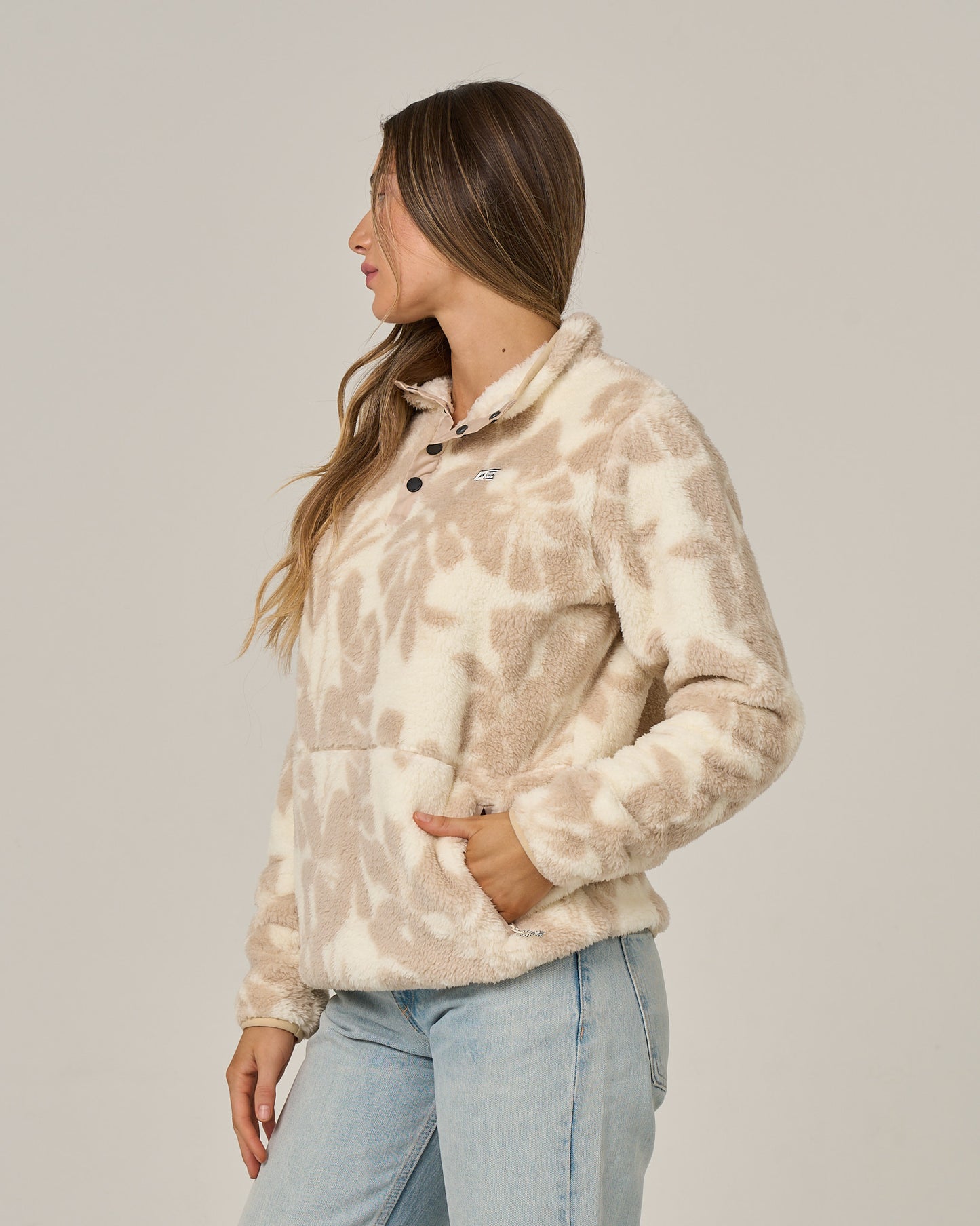 A woman with long brown hair stands in profile against a plain light background, wearing the Salty Crew Calm Seas Pullover in Natural—a beige and white tie-dye sherpa jacket with a mock neck—paired with light blue jeans.