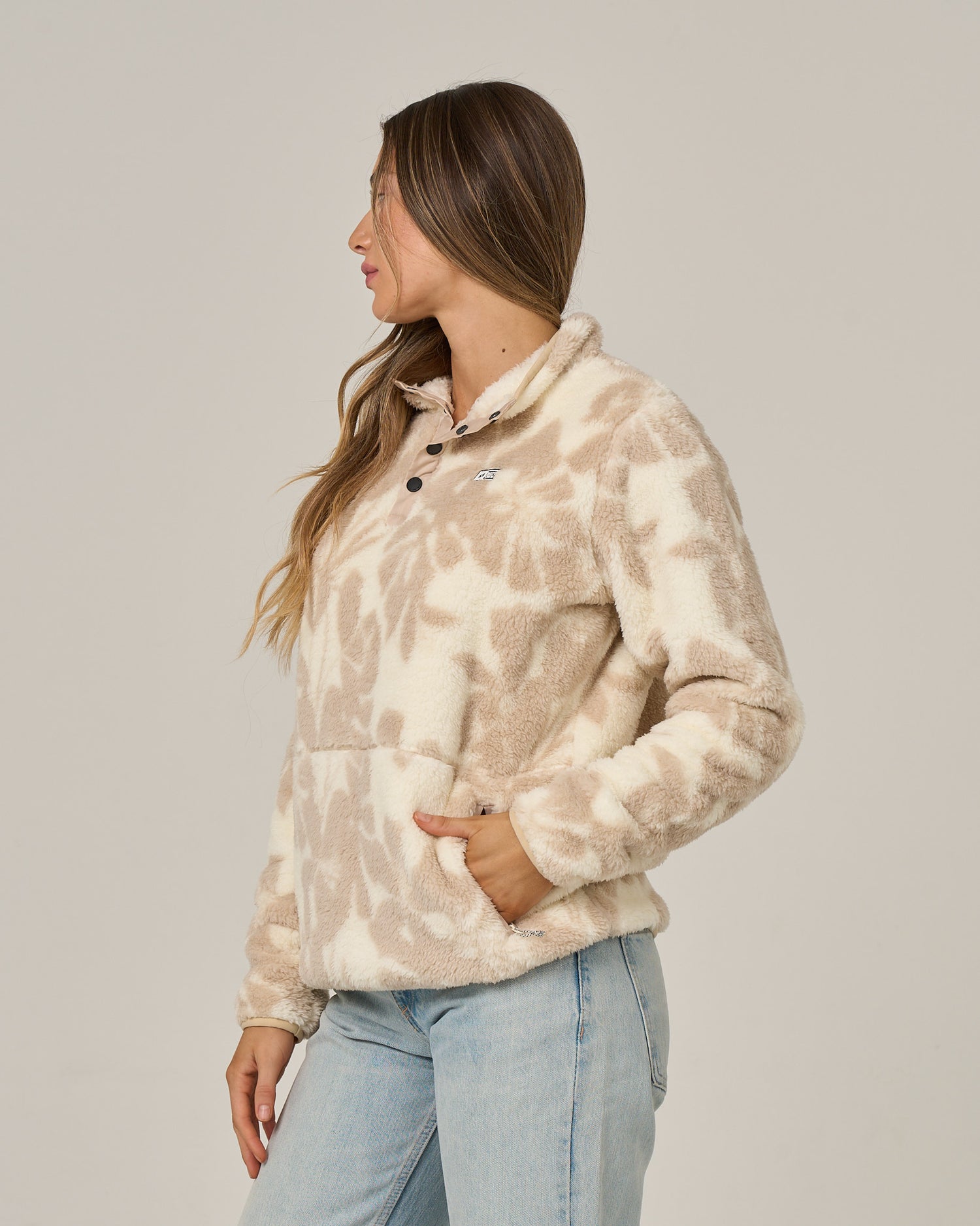 A woman with long brown hair stands in profile against a plain light background, wearing the Salty Crew Calm Seas Pullover in Natural—a beige and white tie-dye sherpa jacket with a mock neck—paired with light blue jeans.