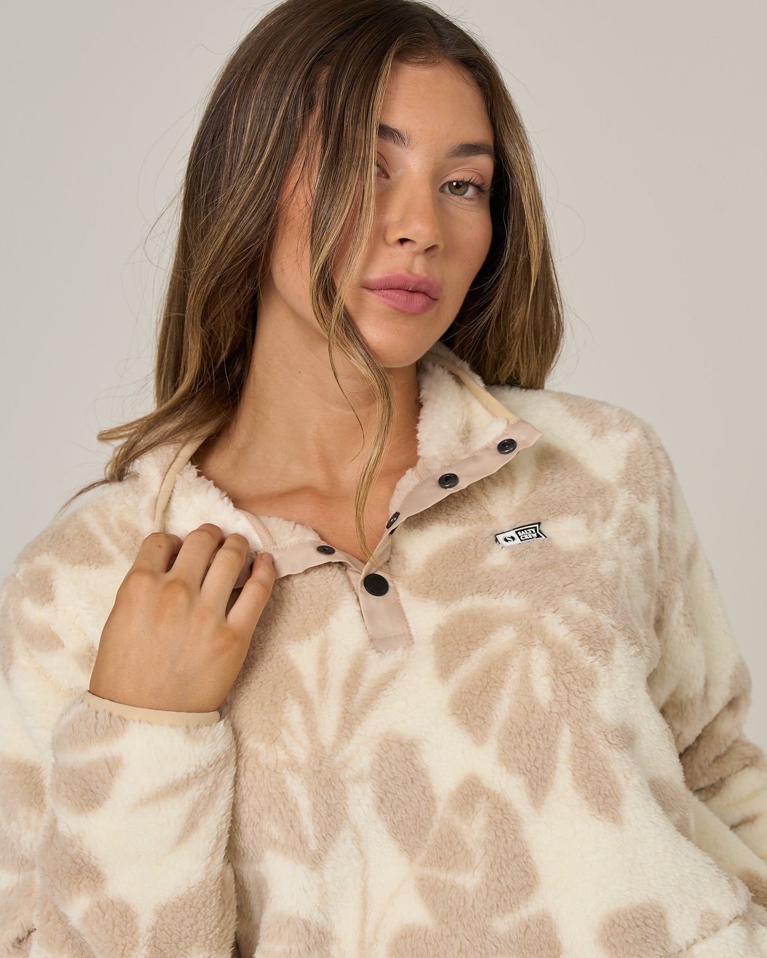 A woman with long hair wears the Salty Crew Calm Seas Pullover in Natural—a cream and beige patterned sherpa fleece featuring a mock neck, black buttons, and a small chest logo—posing with one hand near the collar against a plain background.