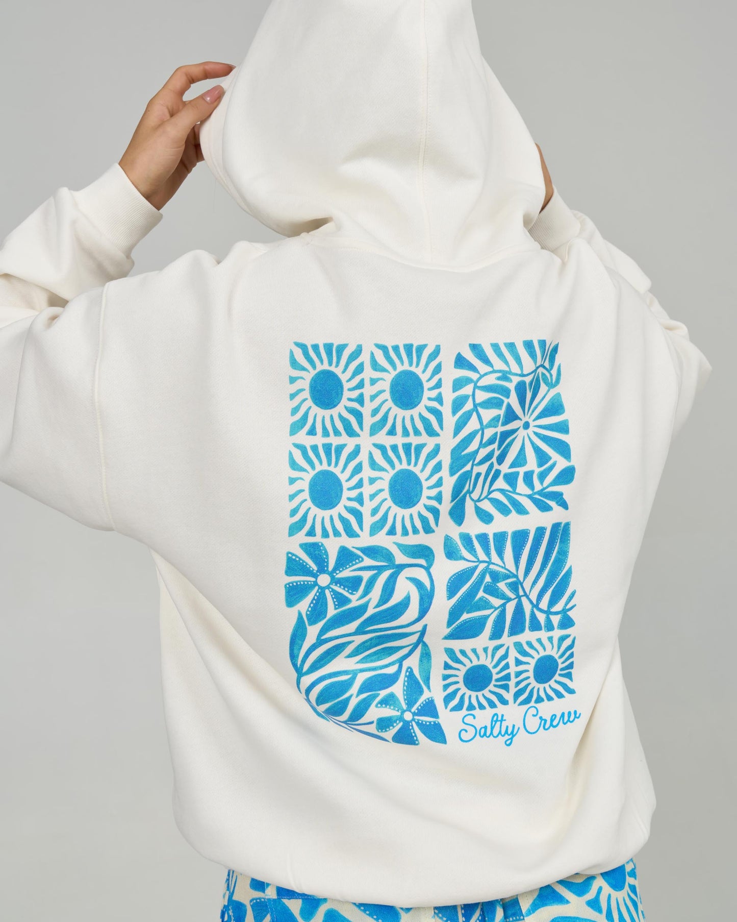 A person adjusts the hood of the Sunblock Hoody - Off White by Salty Crew. The cozy fleece features a blue geometric sun and wave design on the back, classic kangaroo pocket, and Salty Crew branding at the lower right.